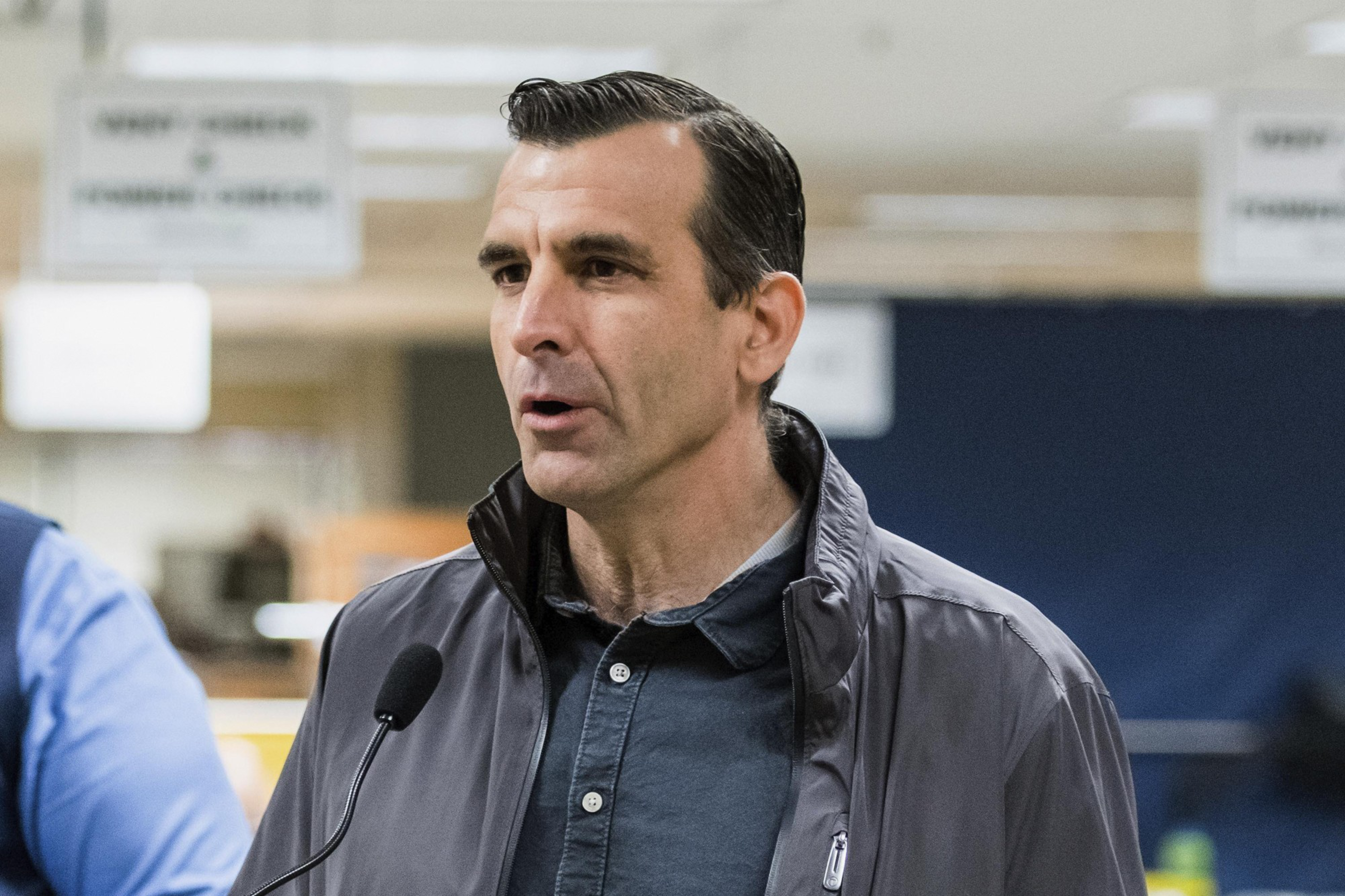 A man in a jacket speaking into a microphone, with blurry office-like background.