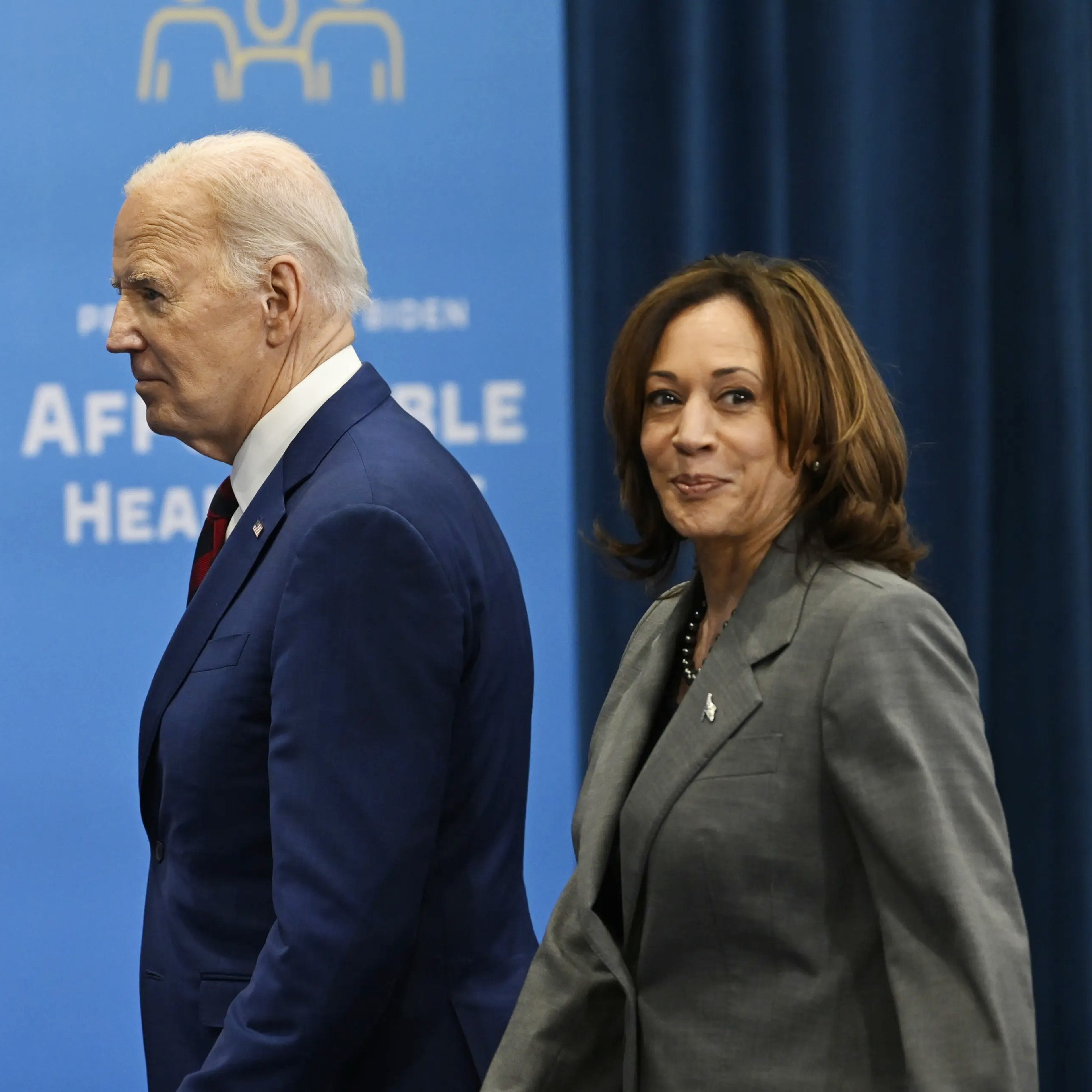 A man in a blue suit and a woman in a gray suit walk in front of a backdrop with the U.S. flag and text promoting affordable healthcare.