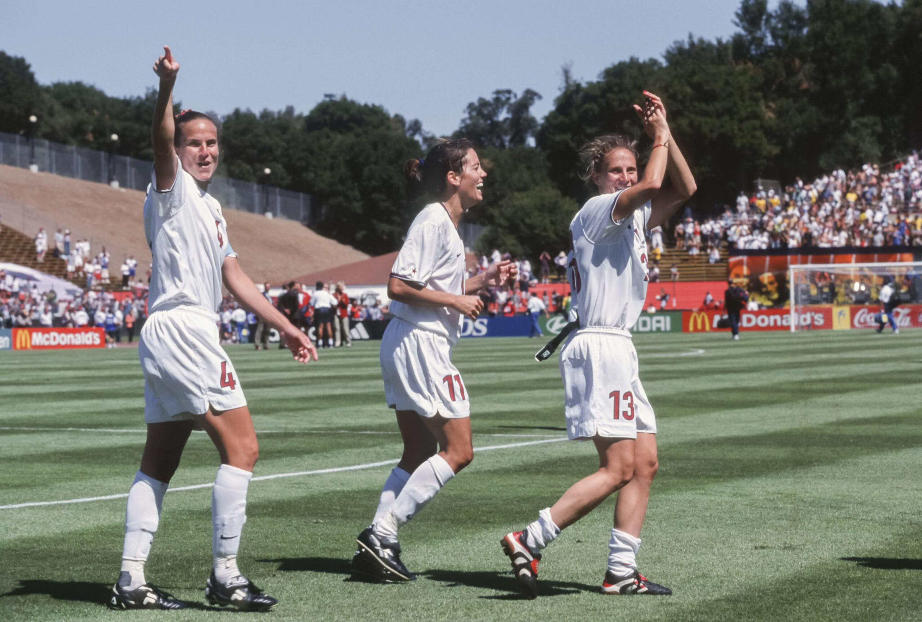 Three female soccer players in white uniforms celebrate on a field, with one pointing, one smiling, and another clapping toward a crowd.