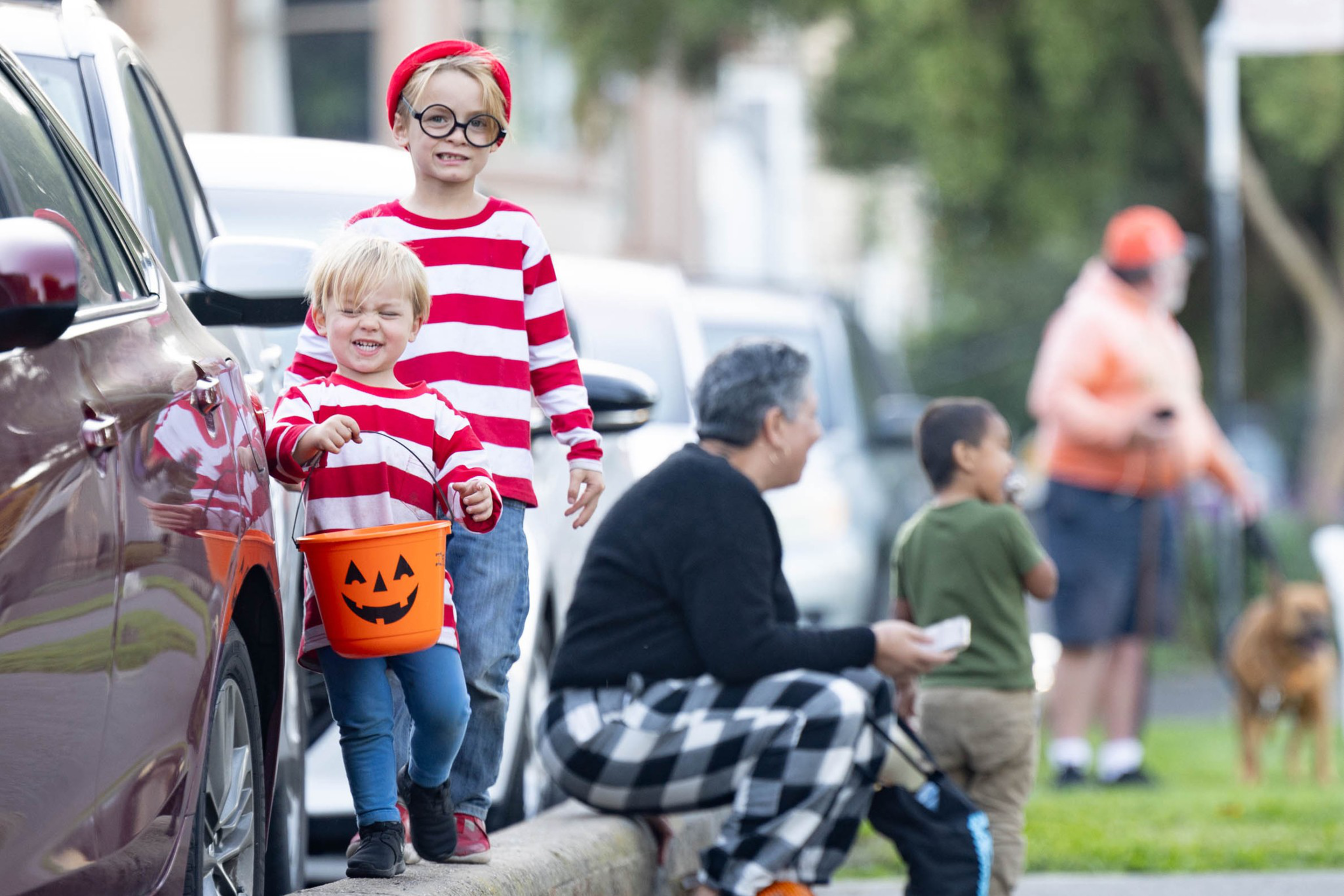 Two kids dressed as Waldo smile while trick-or-treating with an orange pumpkin bucket, as adults and another child watch nearby on a sidewalk.