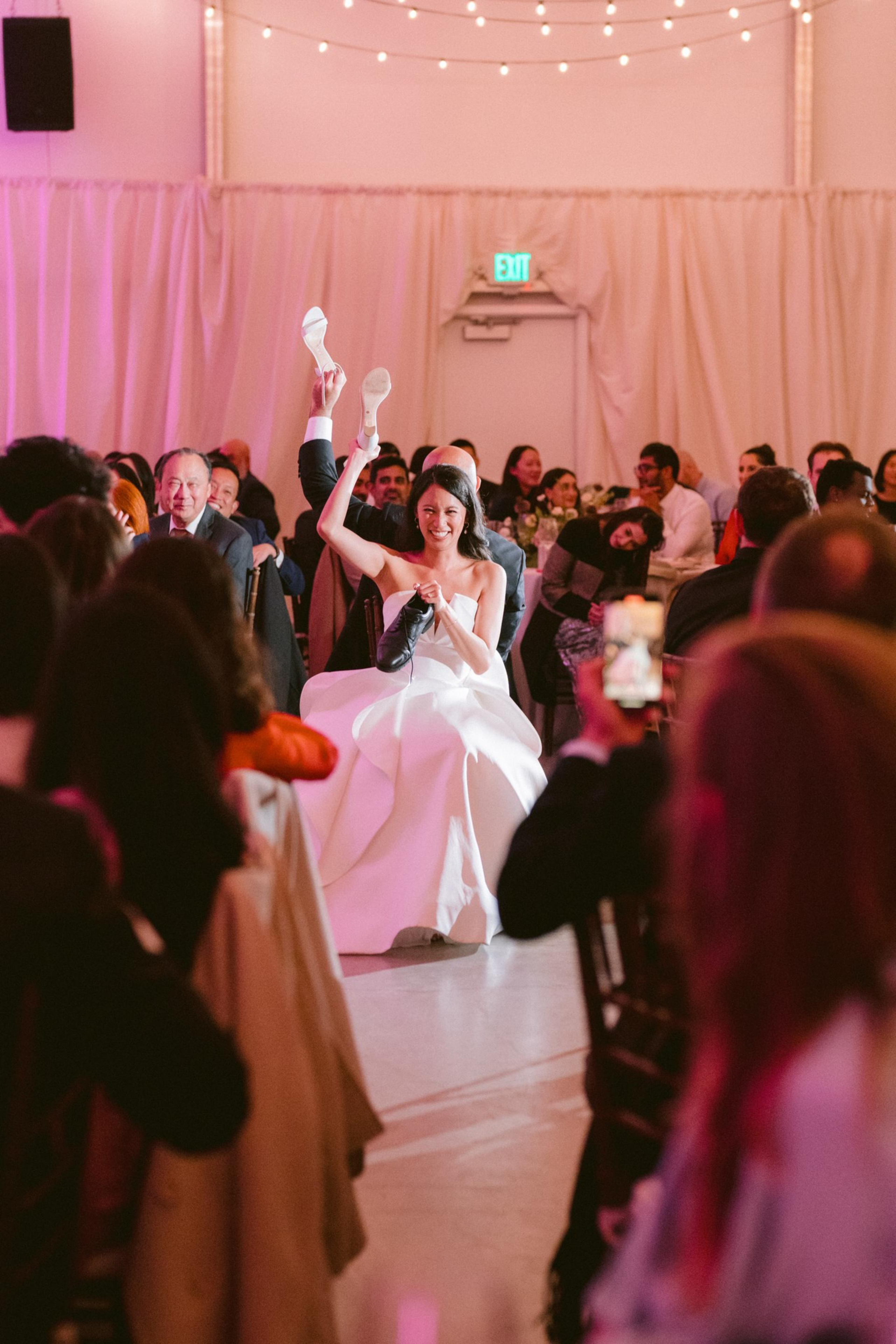 A bride in a white dress sits smiling, holding up two shoes—one white and one black—while wedding guests watch and laugh around her.