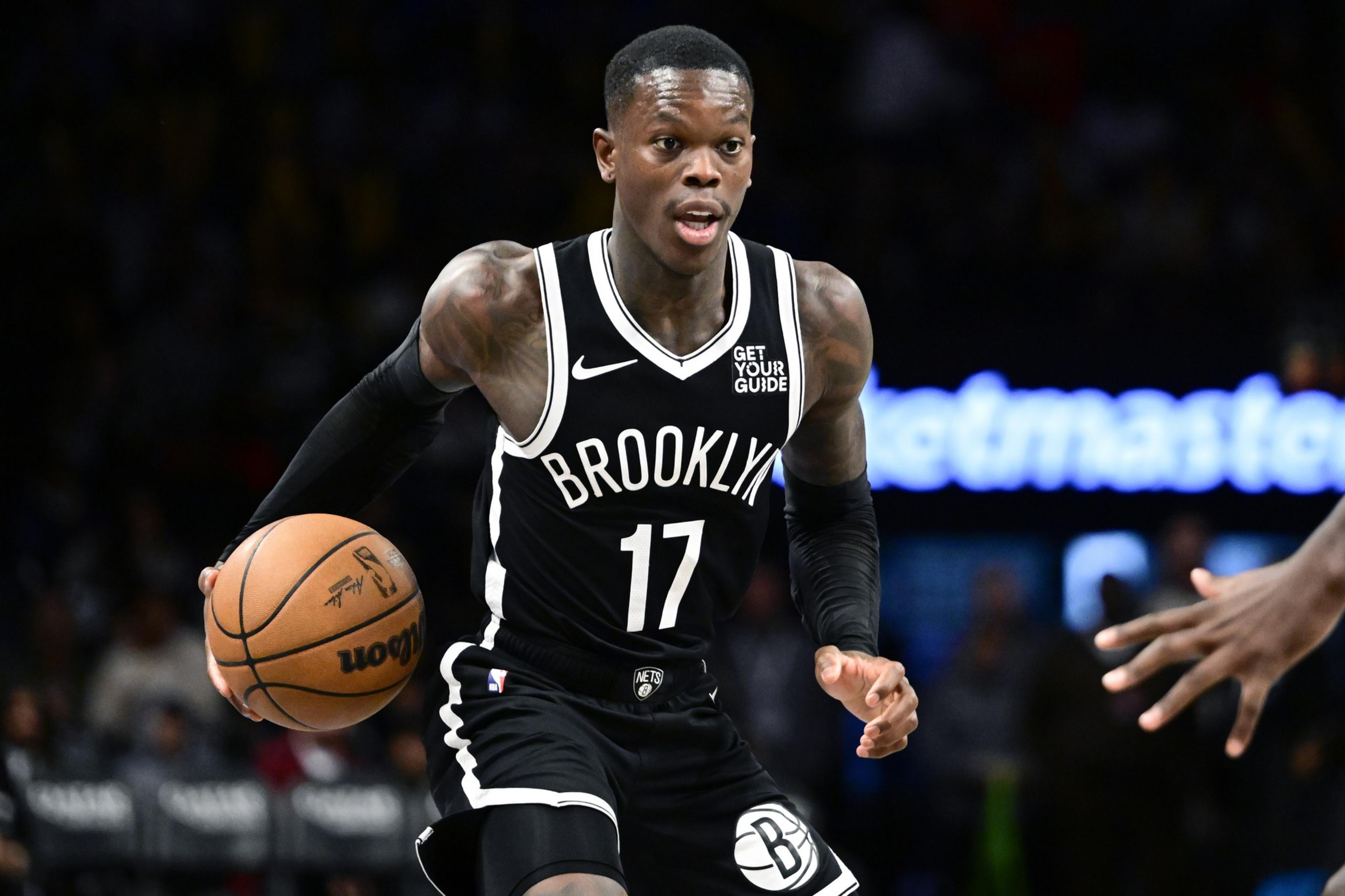 A basketball player in a black Brooklyn jersey, number 17, dribbles the ball on a court. He's focused and in action, with a blurred audience in the background.