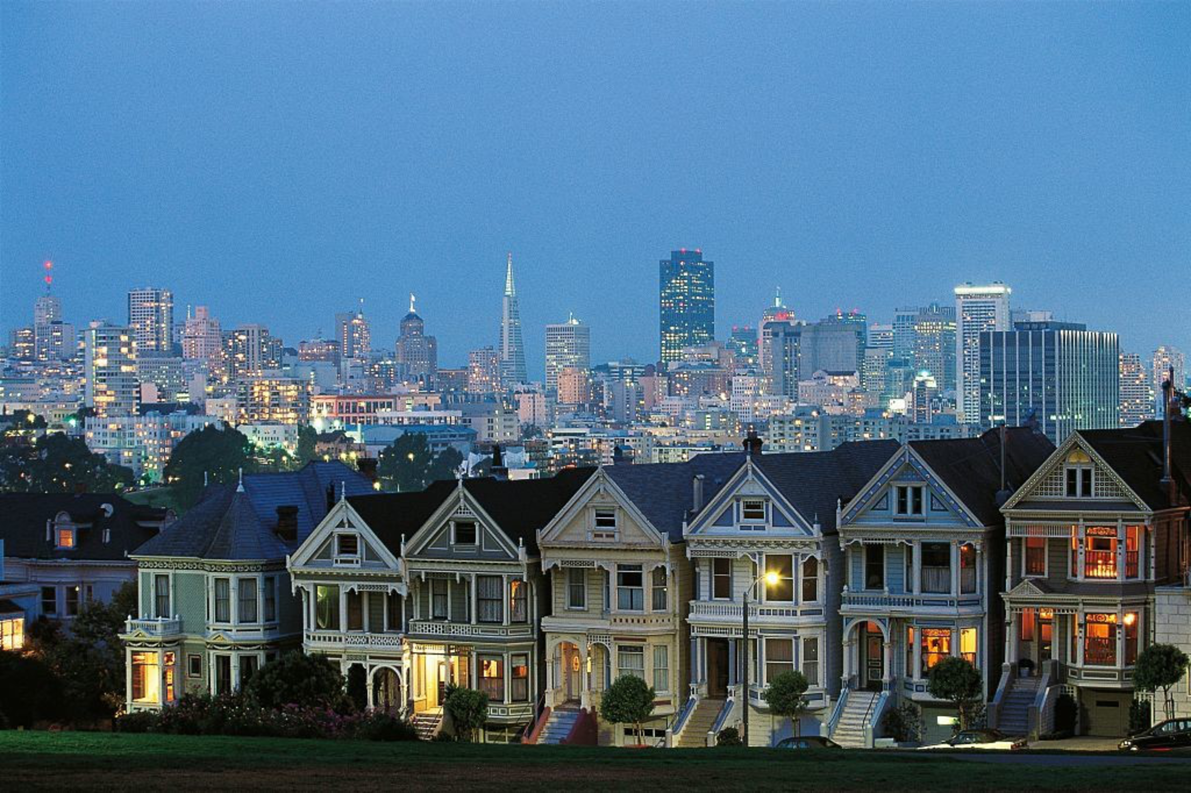 A row of Victorian homes stand in a row with San Francisco's skyline in the background.
