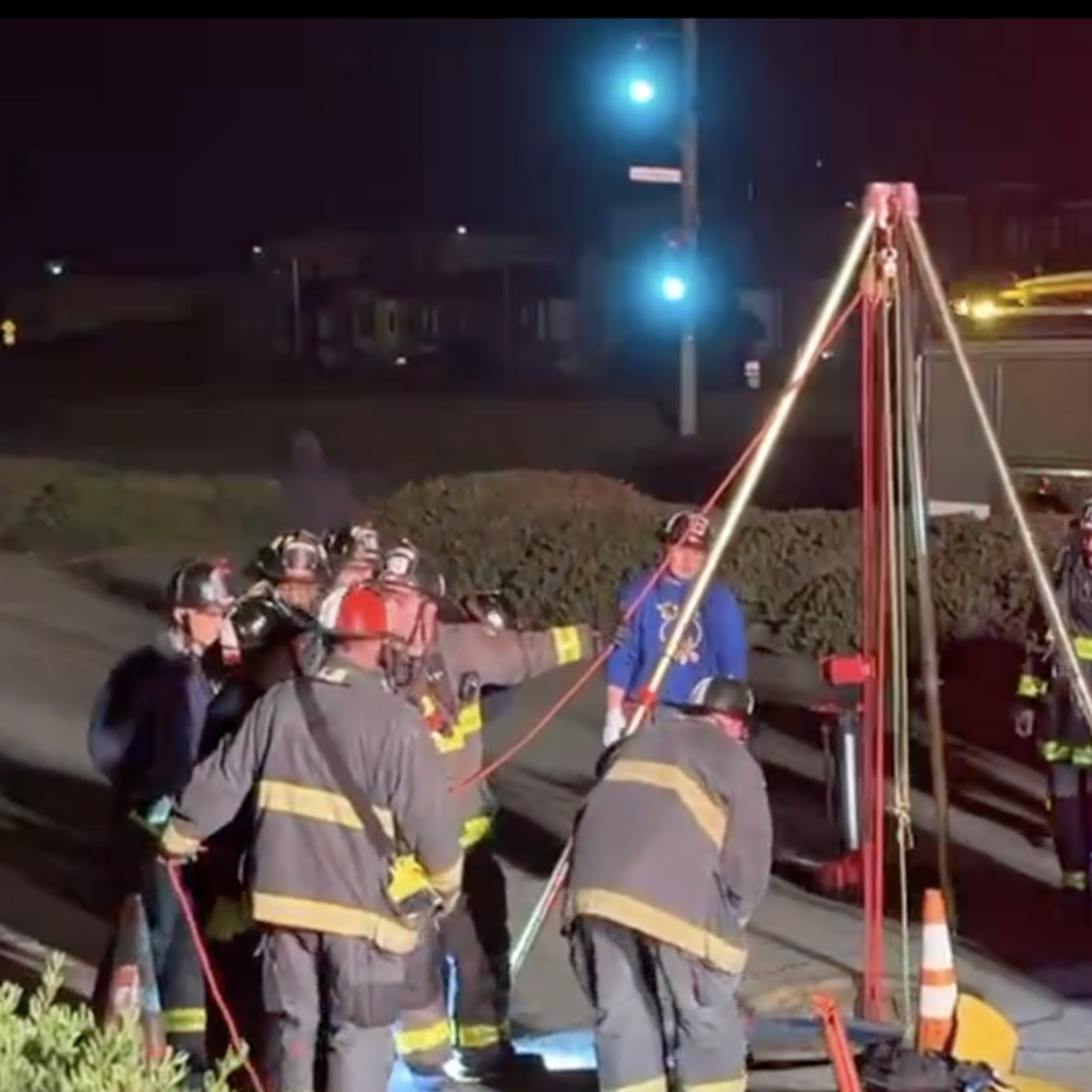 Firefighters are gathered around a tripod setup with ropes and equipment on the roadside at night, illuminated by emergency vehicle lights in the background.