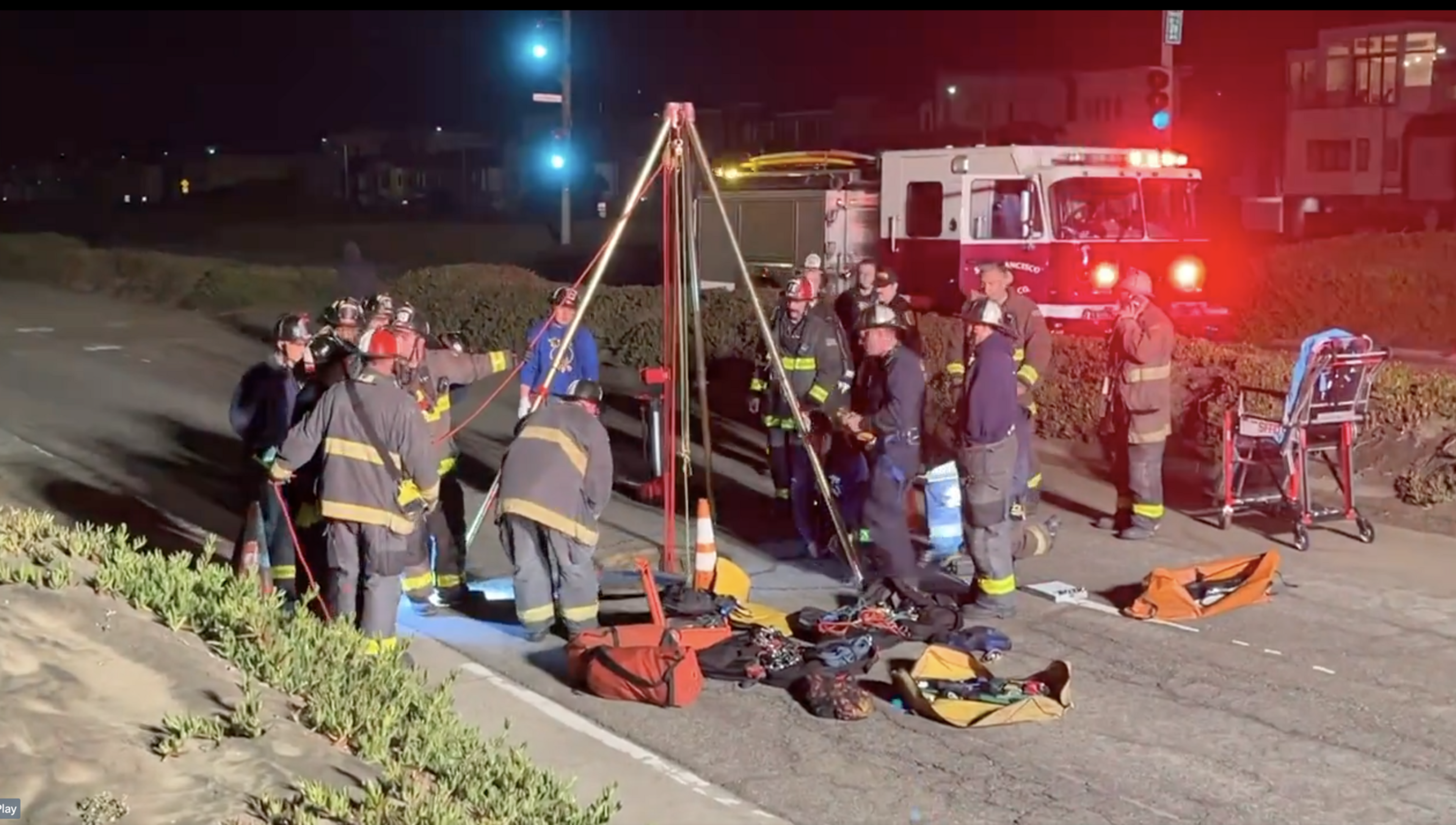 Firefighters are gathered around a tripod setup with ropes and equipment on the roadside at night, illuminated by emergency vehicle lights in the background.