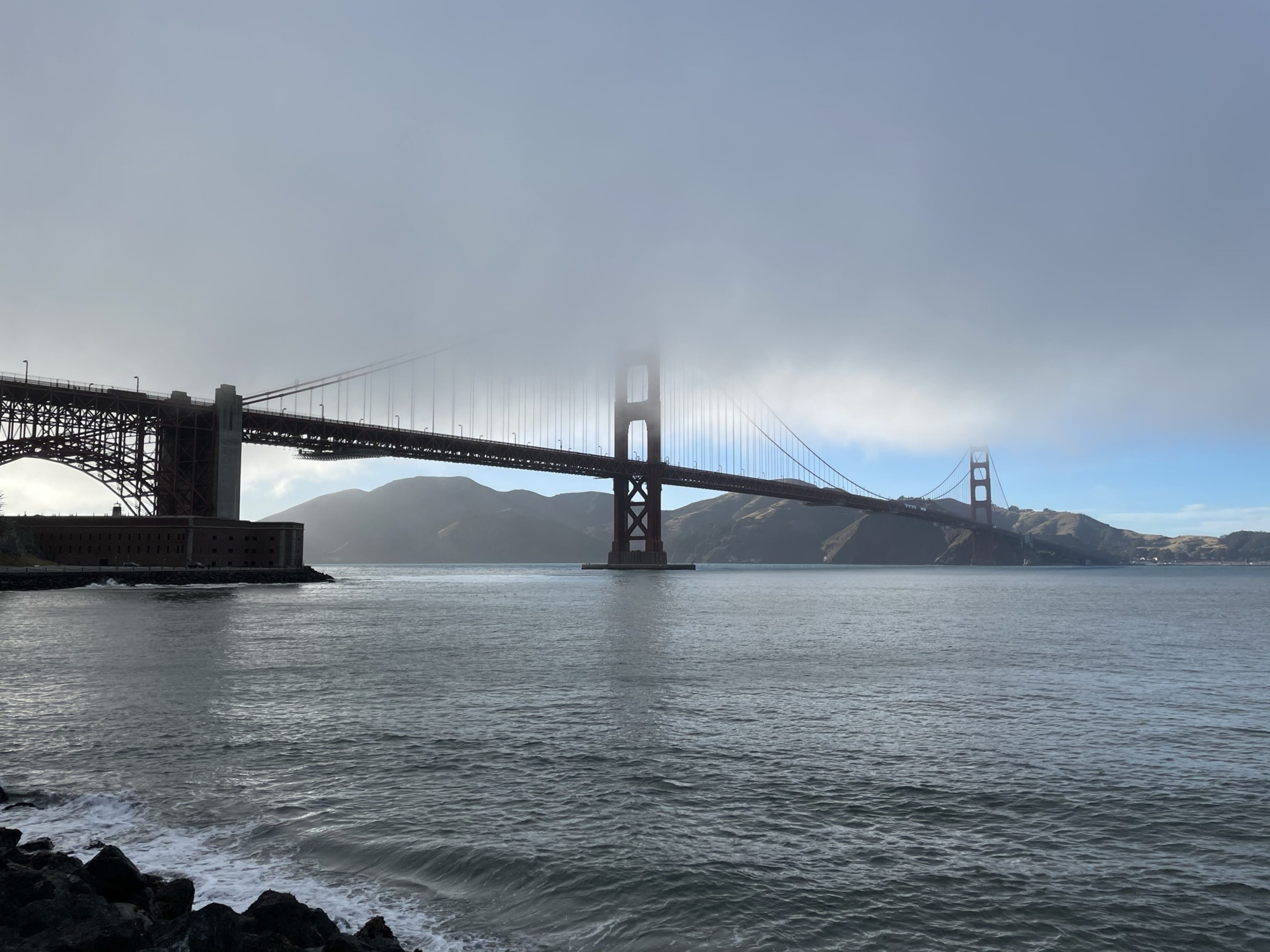 A large suspension bridge spans a calm body of water, partially shrouded in fog. In the background, there are hills under a mostly clear sky.