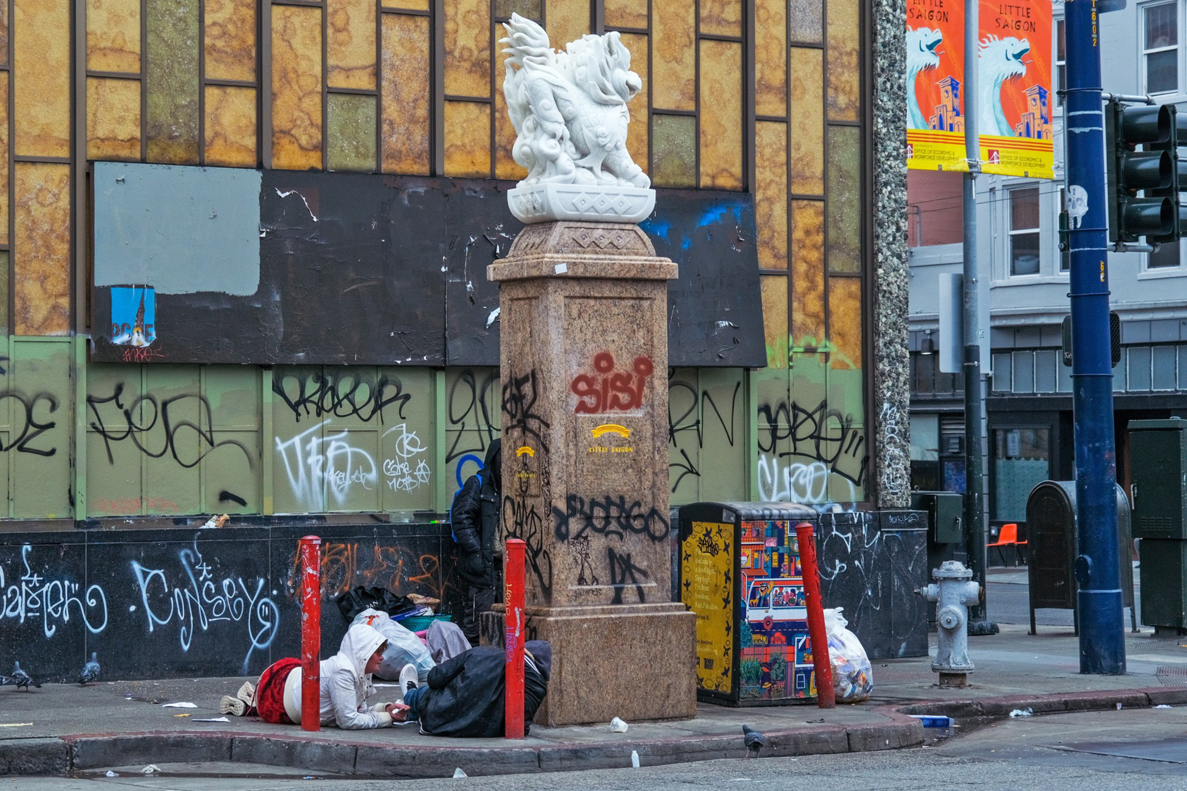 A graffiti-laden urban corner with a person resting by a statue base, surrounded by trash and discarded items.