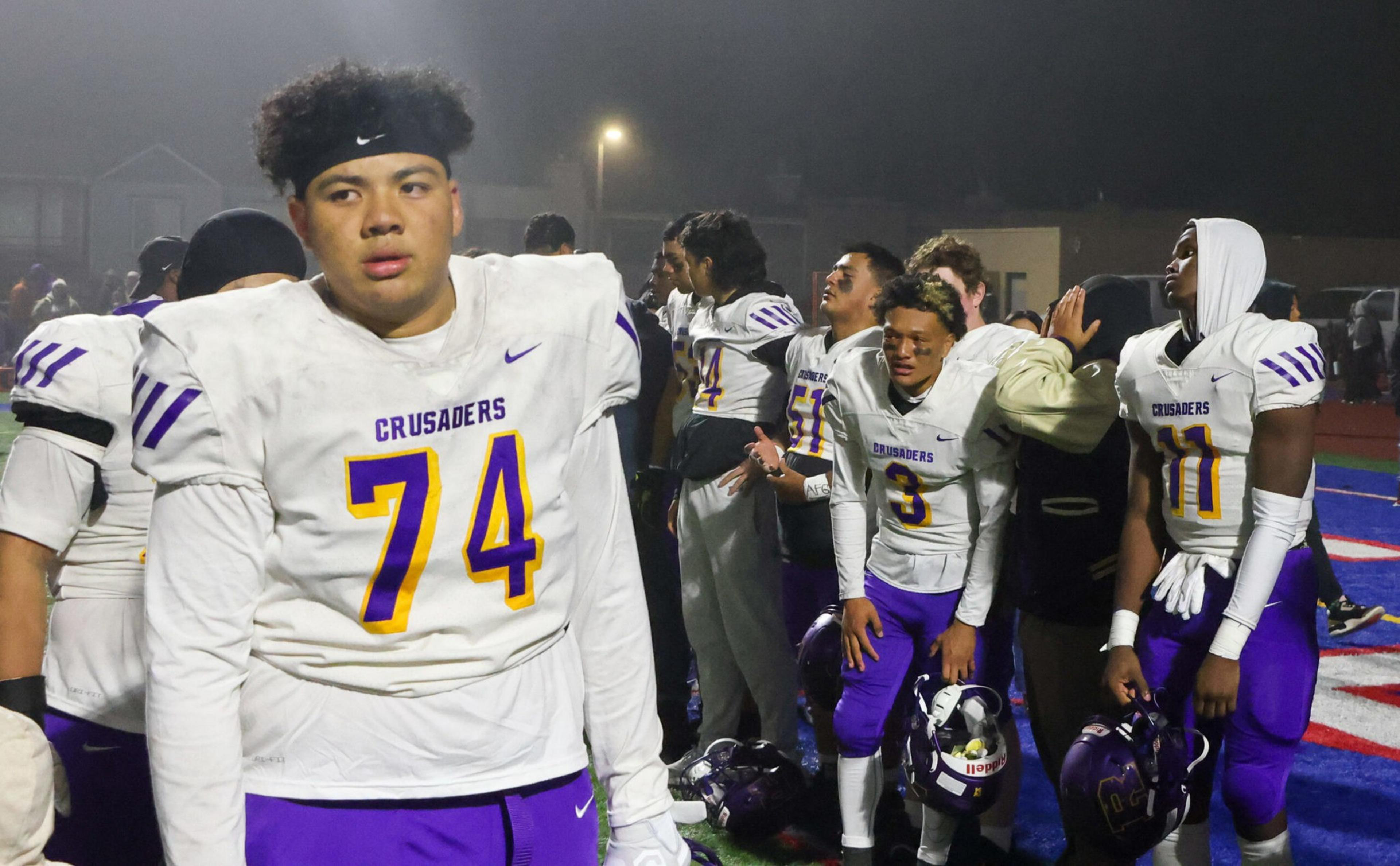 Several football players in white jerseys and purple pants stand on a field at night, looking tired and holding their helmets.