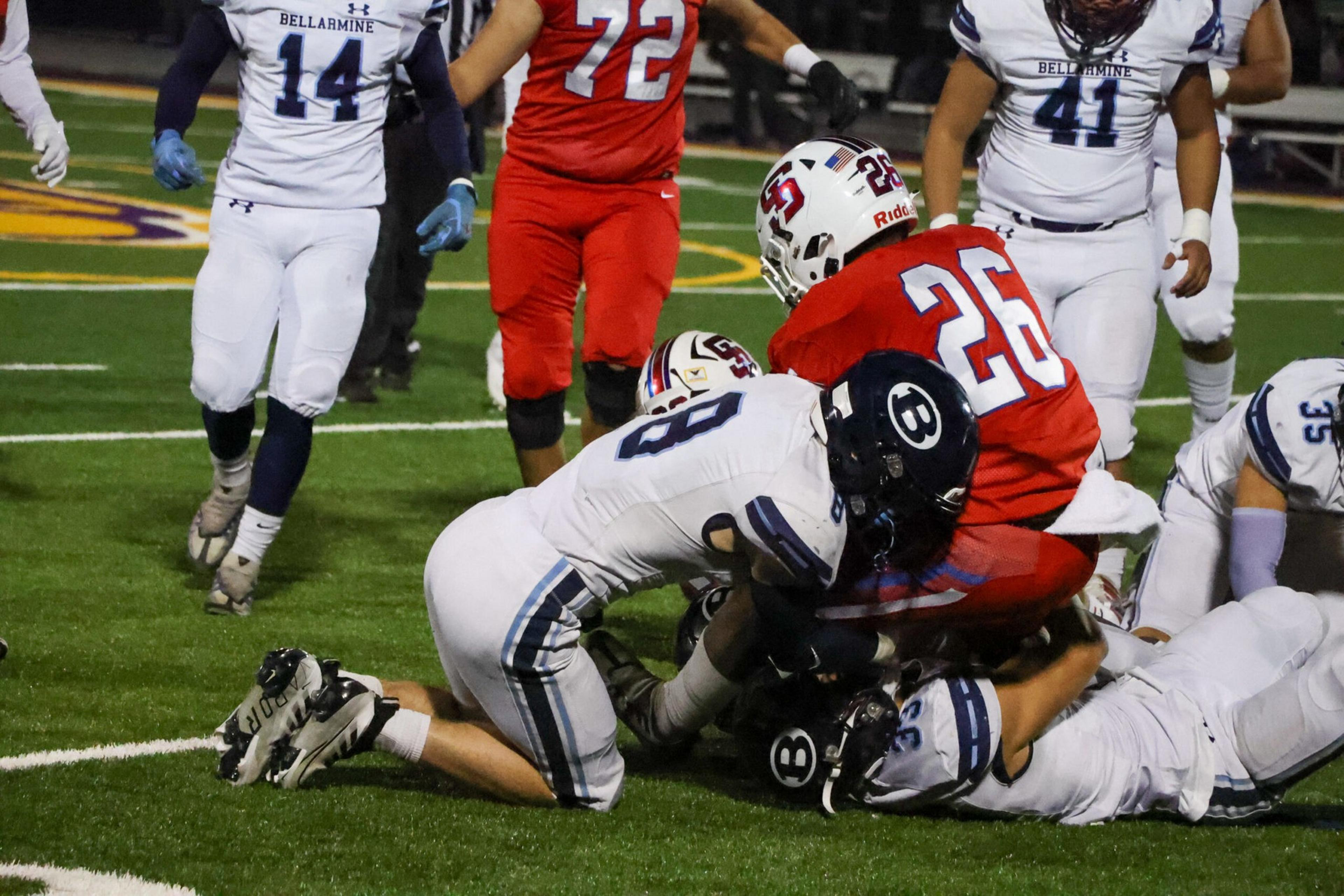 A football player in red is tackled to the ground by two players in white uniforms with dark helmets on a green field.