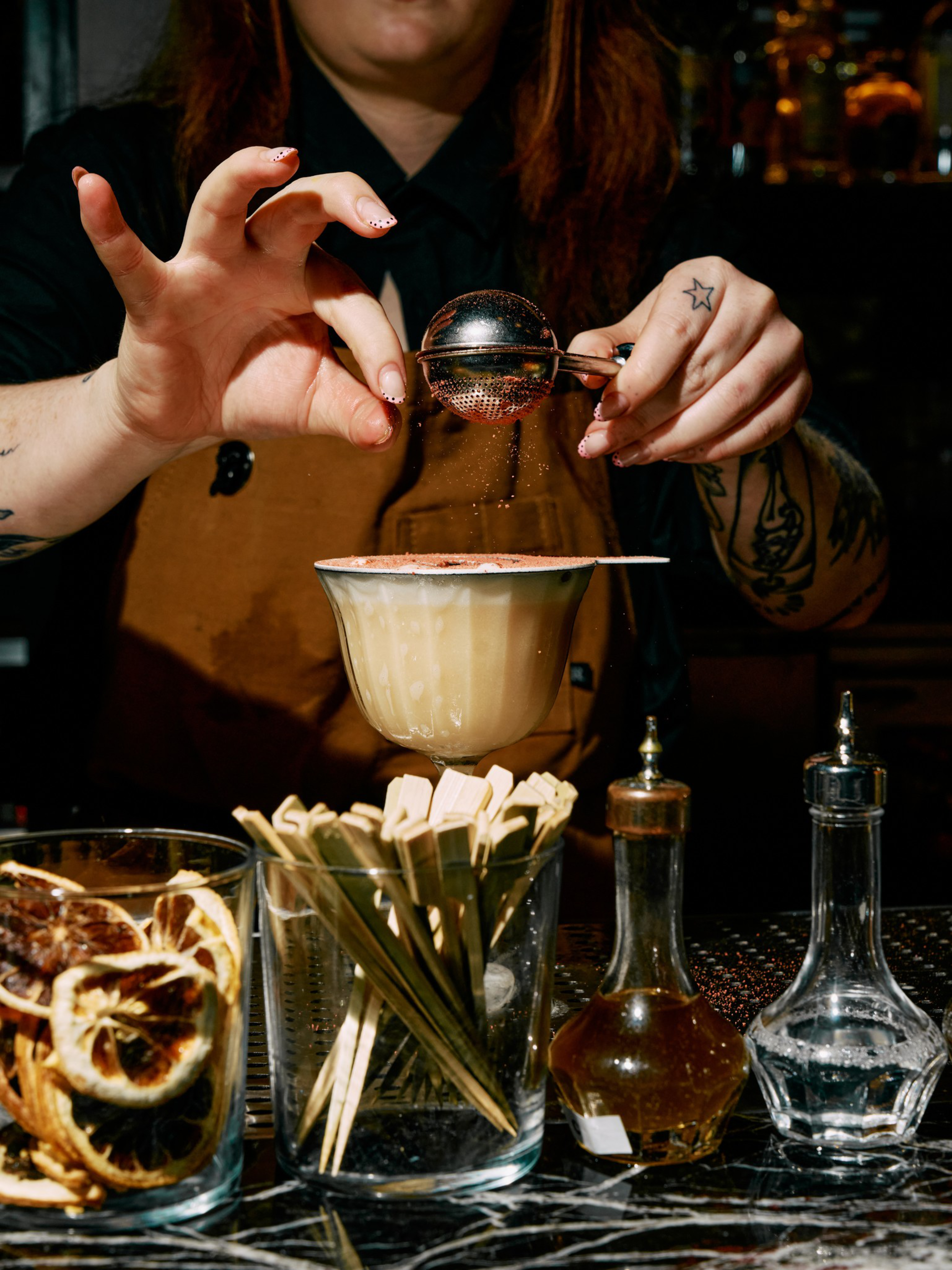 A person sprinkles powder over a creamy cocktail in a coupe glass, with dried citrus, stirrers, and bottles on a marble bar counter.