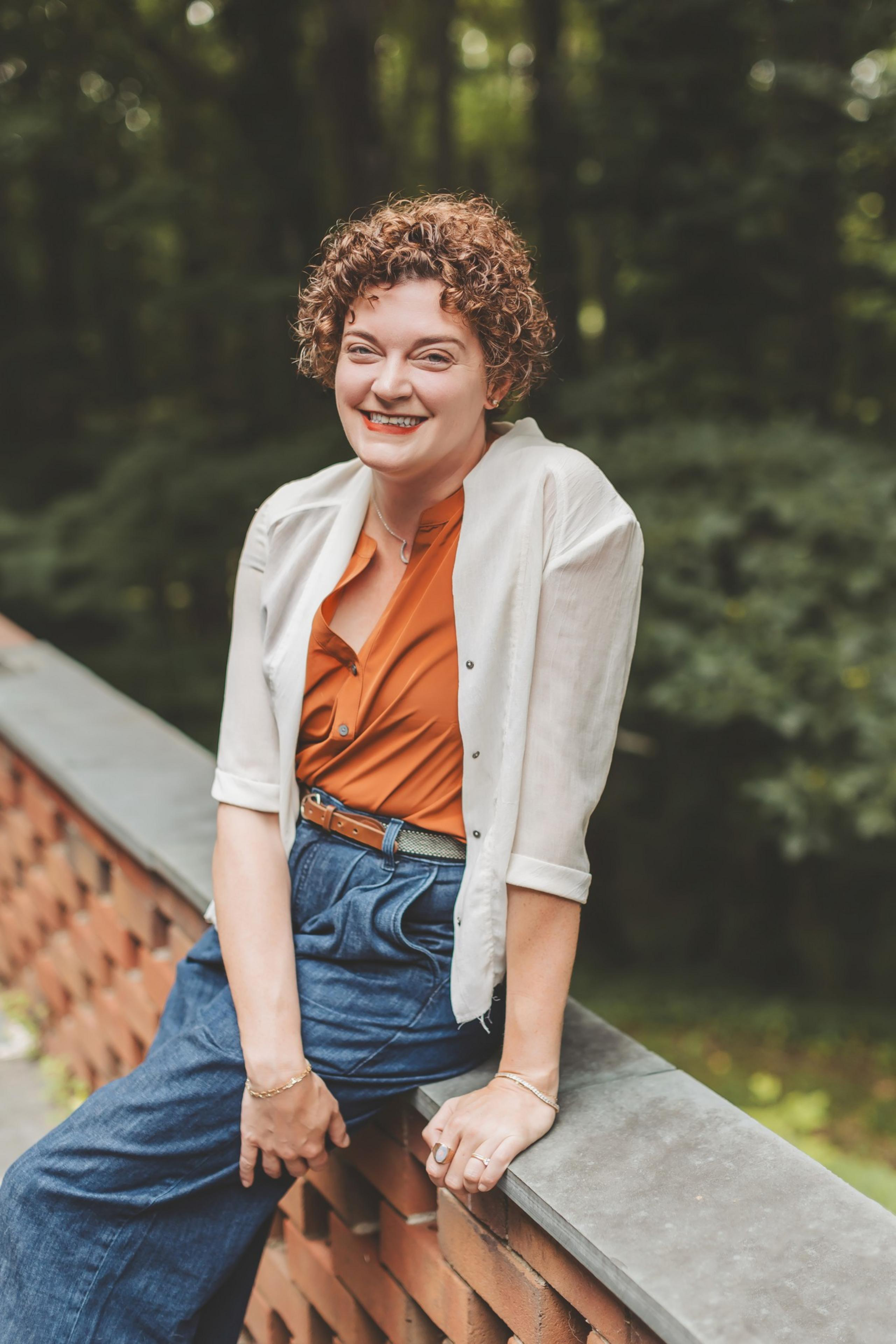 A smiling woman with curly hair, wearing an orange shirt, white cardigan, and blue pants, leans on a brick and stone railing with greenery in the background.