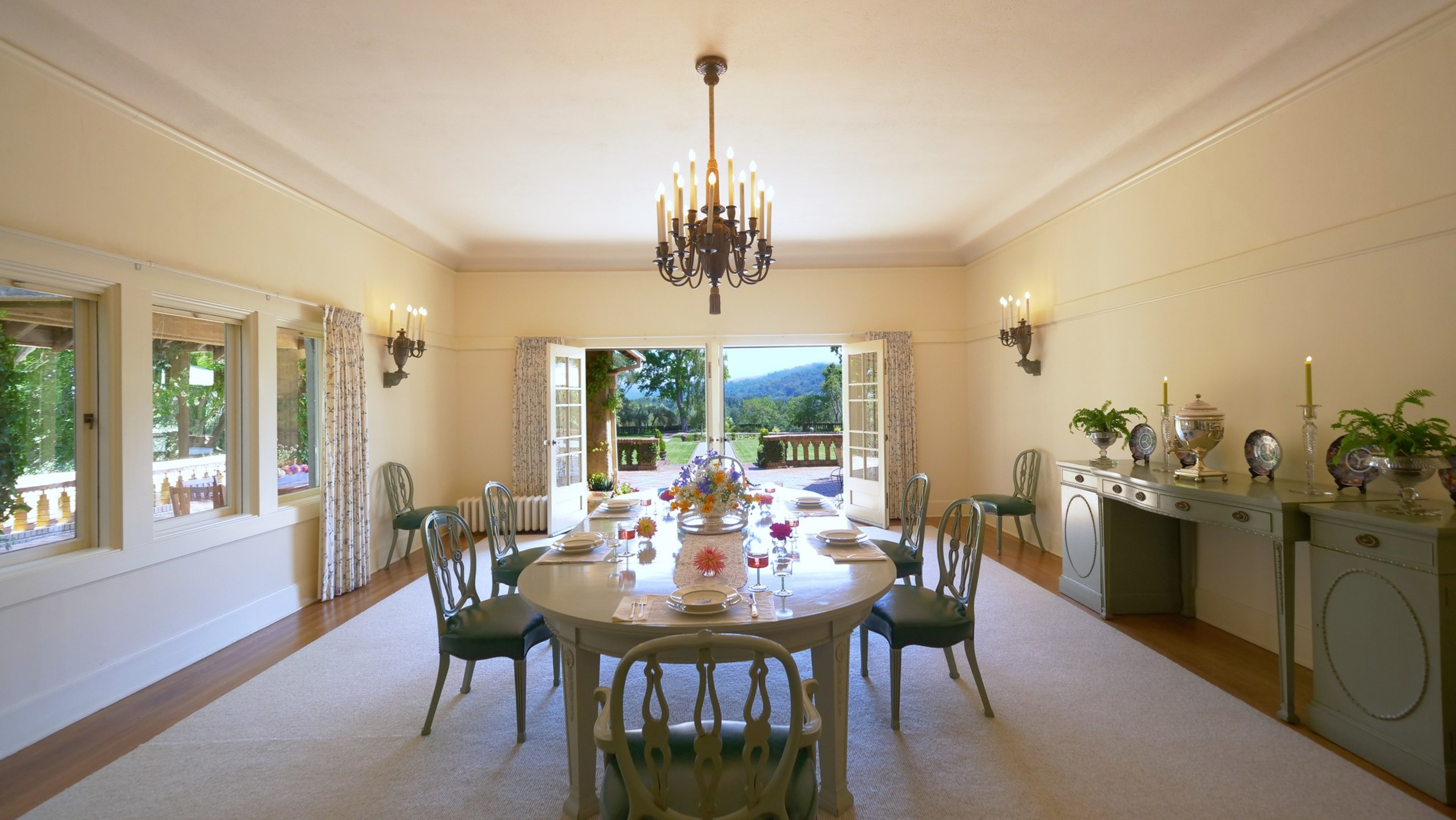 A bright dining room features a large oval table set with plates, glasses, flowers, eight chairs, a chandelier, and double doors opening to a garden view.