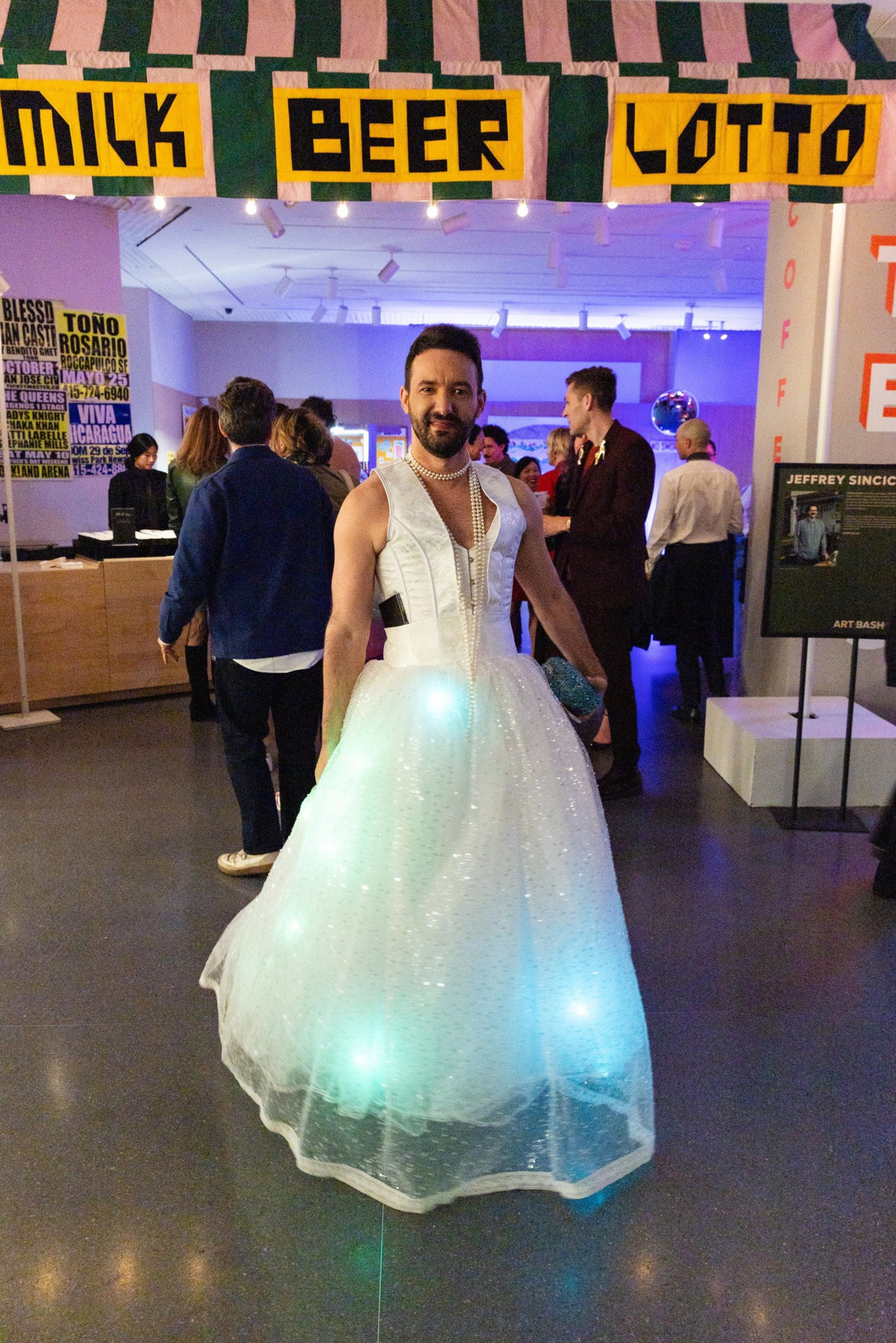 A person in a lit-up, sparkly white gown stands smiling at an event. Behind them, a colorful sign reads &quot;MILK BEER LOTTO,&quot; and people are gathered in conversation.