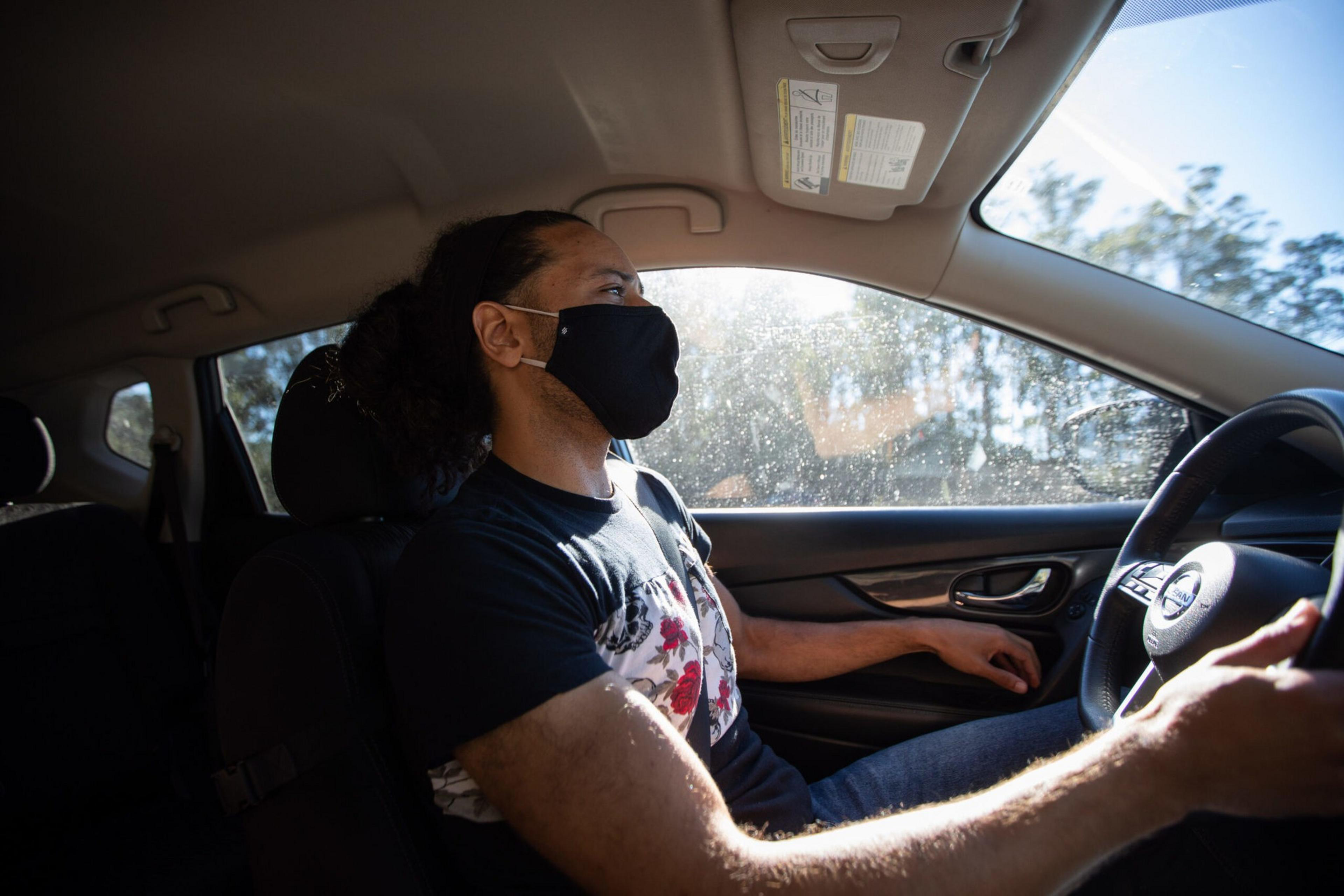 A man wearing a black face mask and a graphic t-shirt is driving a car with sunlight streaming through the window, gripping the steering wheel with his right hand.
