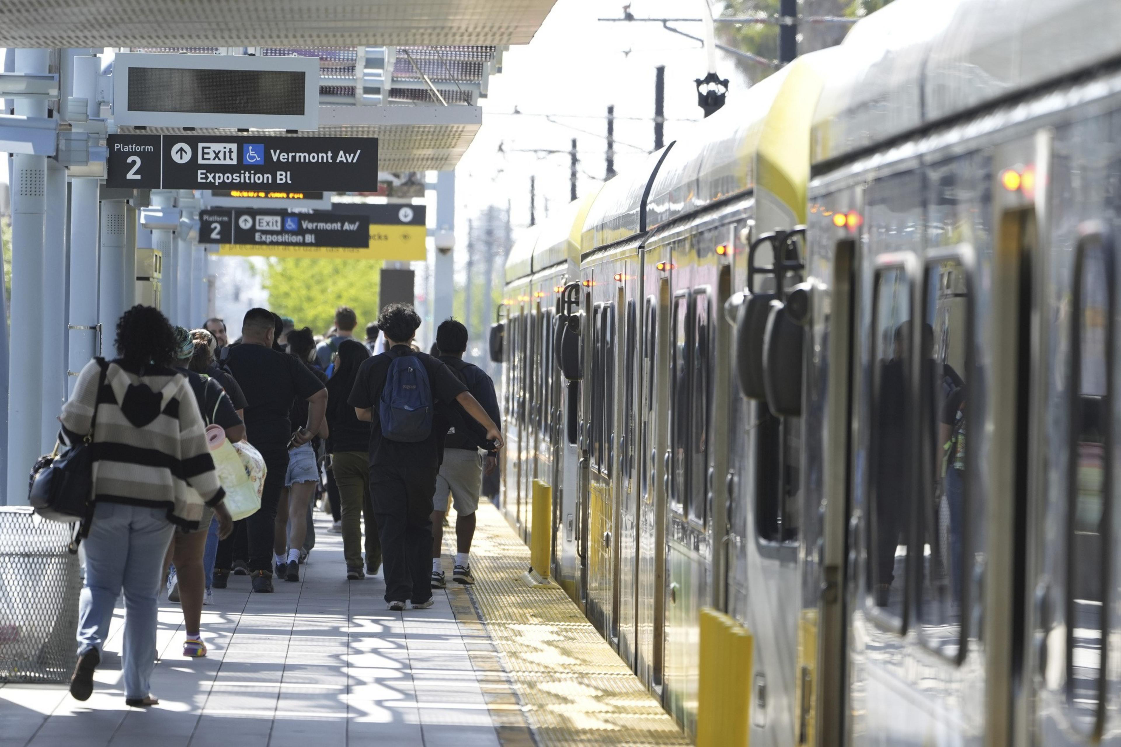 Passengers are walking on a train platform alongside a yellow train. Signs for exits and platform numbers are above, with people heading towards the exit.