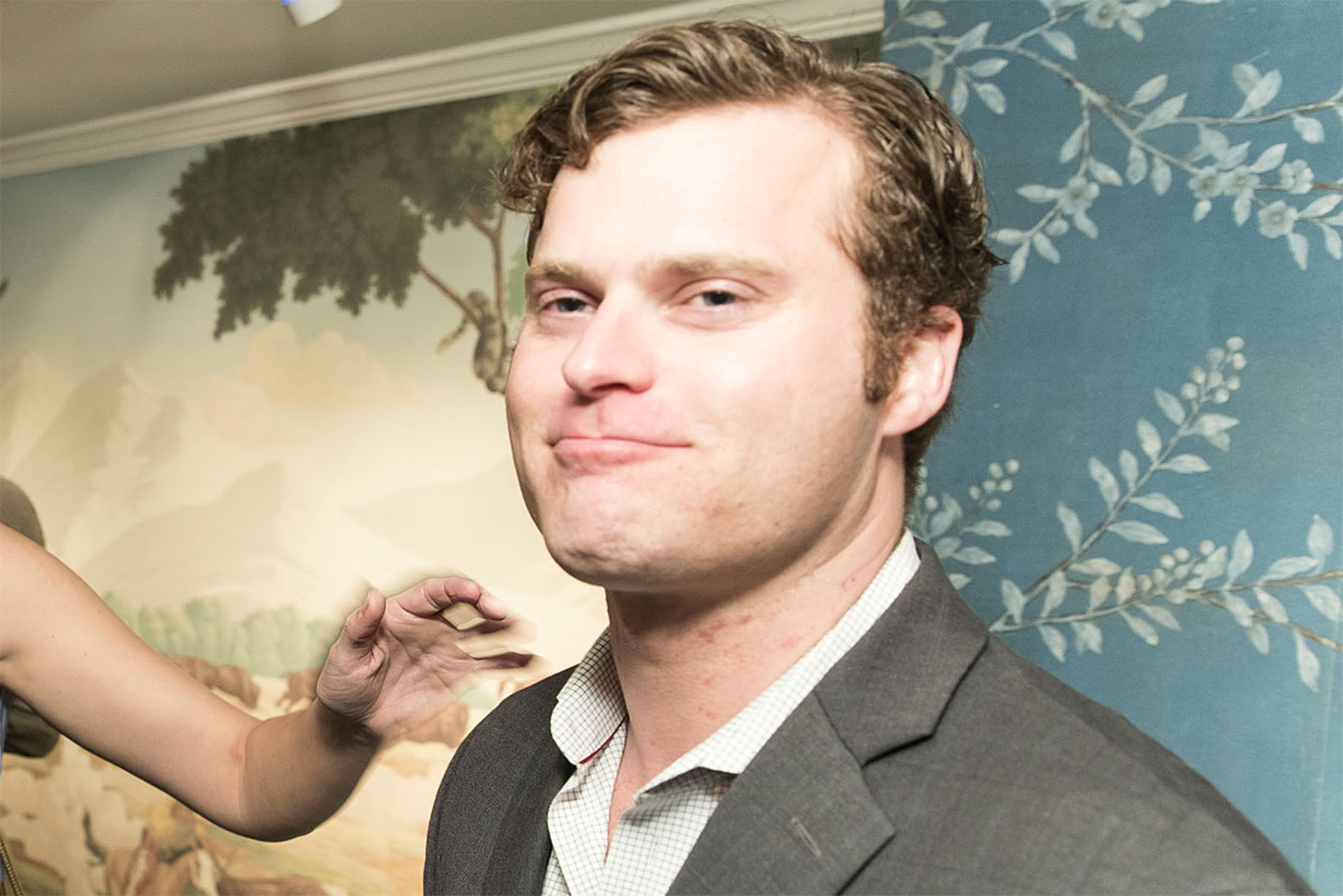 A man in a suit smirks while someone's hand makes a playful gesture near his face.