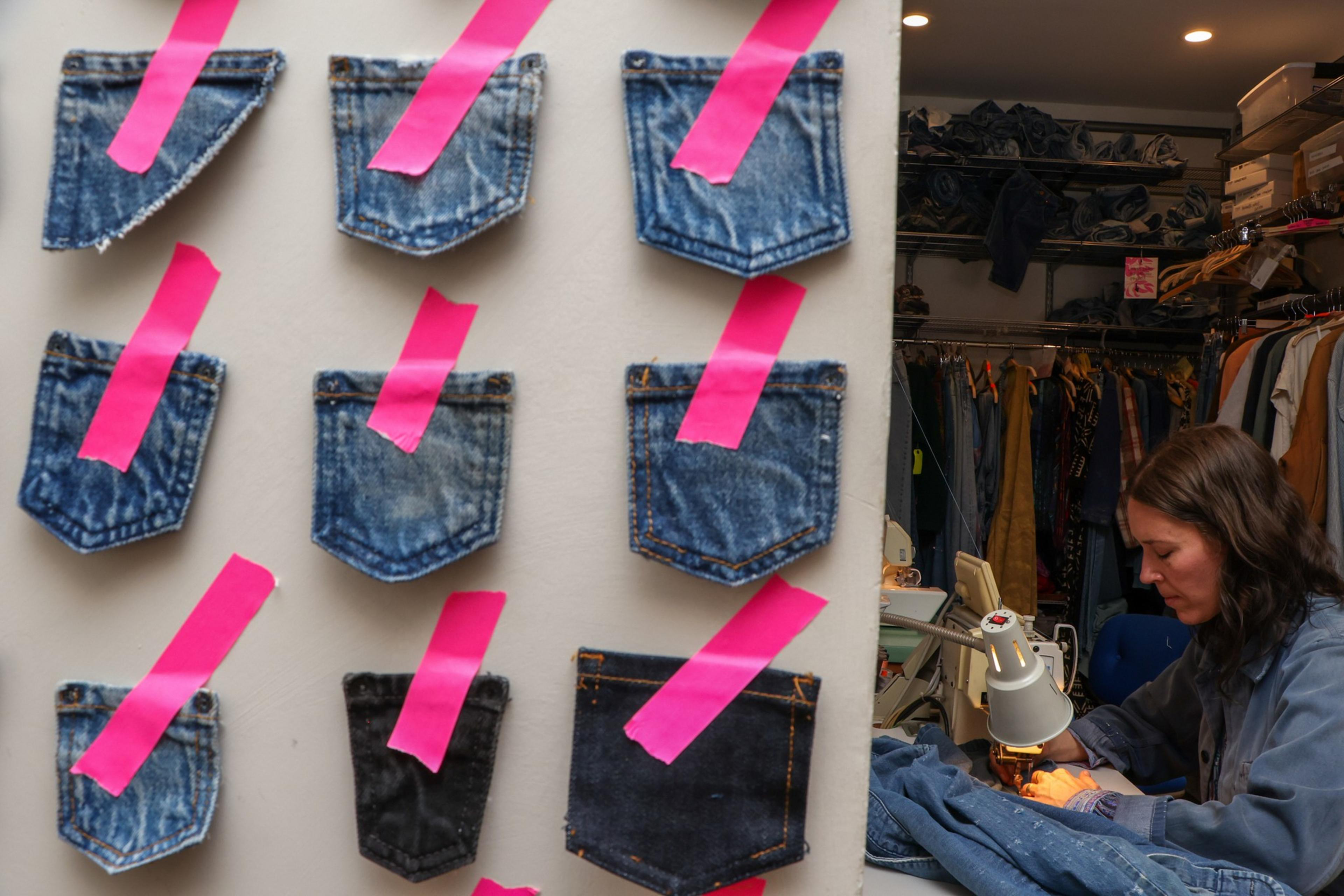 Nine denim jean pockets are taped to a wall with pink tape, while a woman sews fabric at a machine in a cluttered workspace.