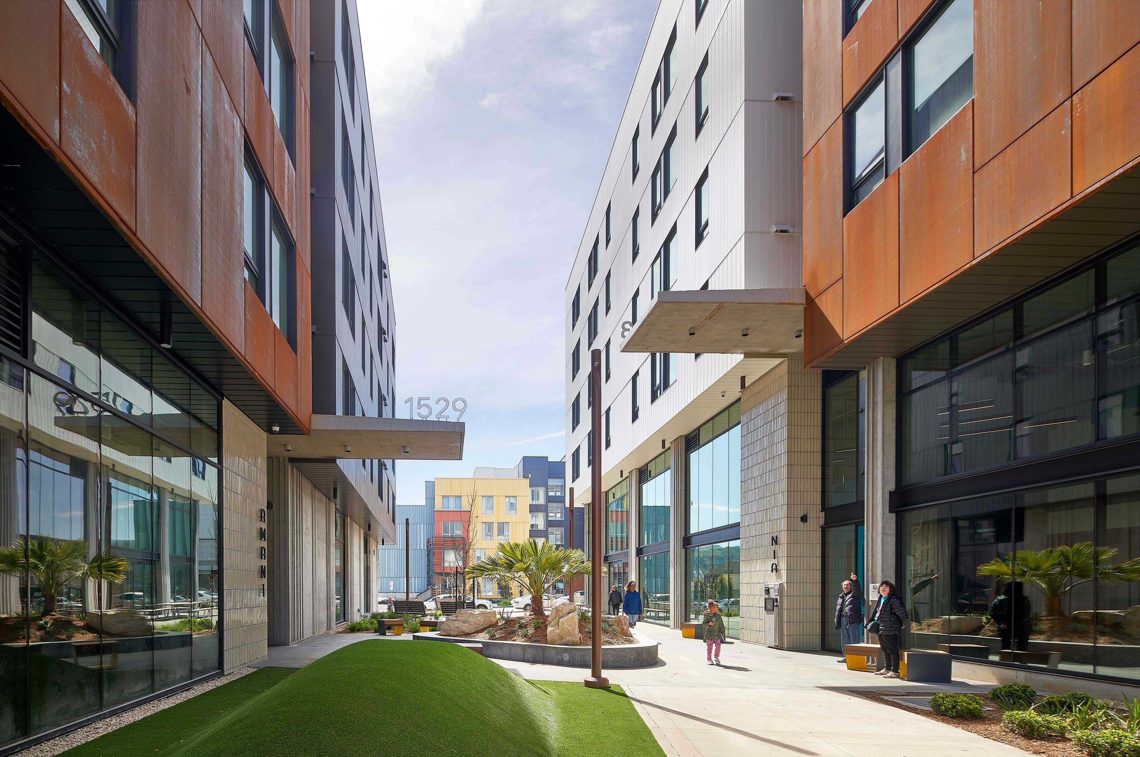Two modern buildings with large windows border a landscaped plaza with greenery, rocks, palm trees, and a few people walking and sitting.