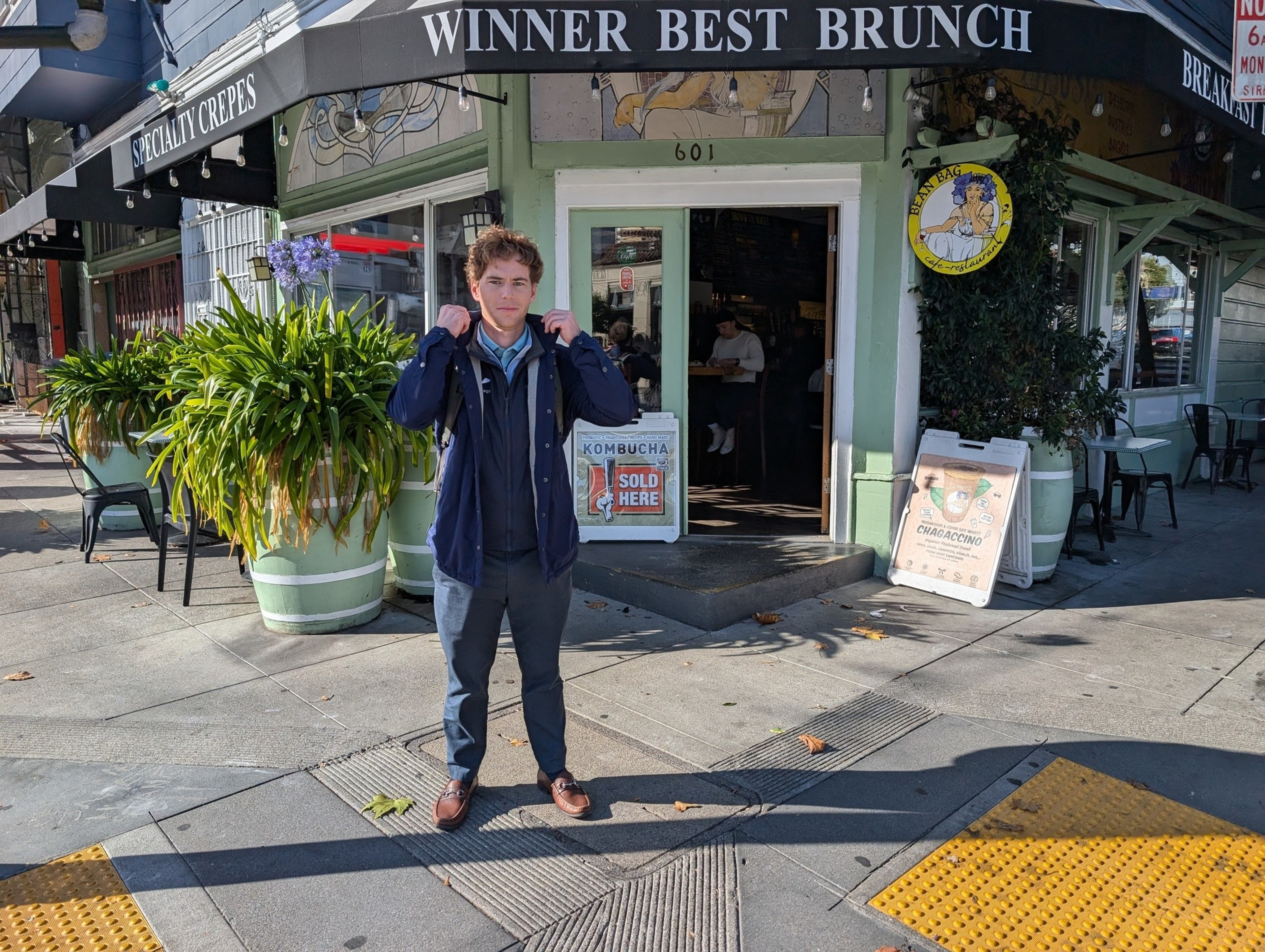 A person stands outside a cafe with a "Winner Best Brunch" sign. They're wearing a jacket and have a backpack. There are large potted plants and signs nearby.