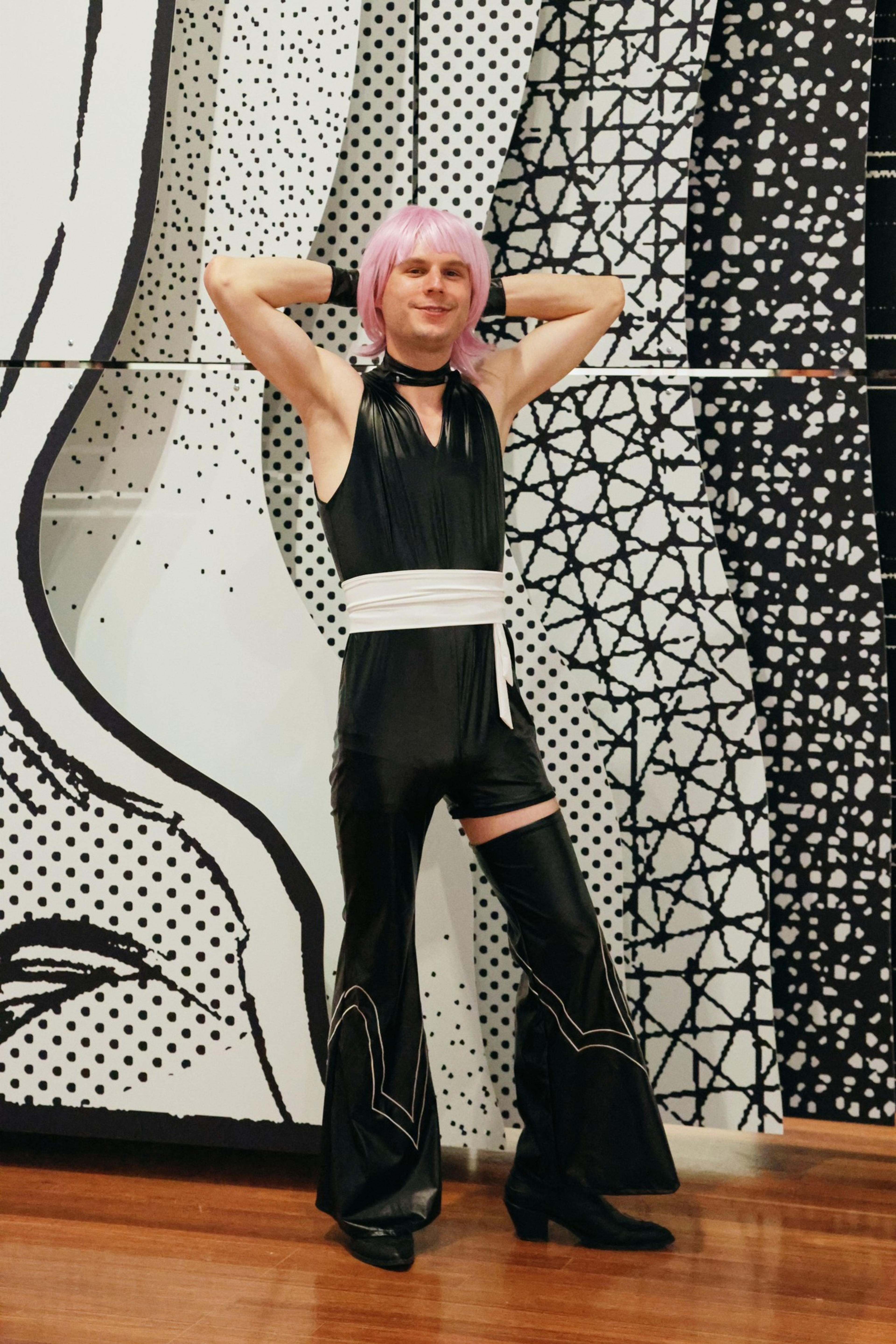 A person with pink hair wears a black sleeveless jumpsuit, white belt, and black boots, posing confidently against a black-and-white patterned wall.