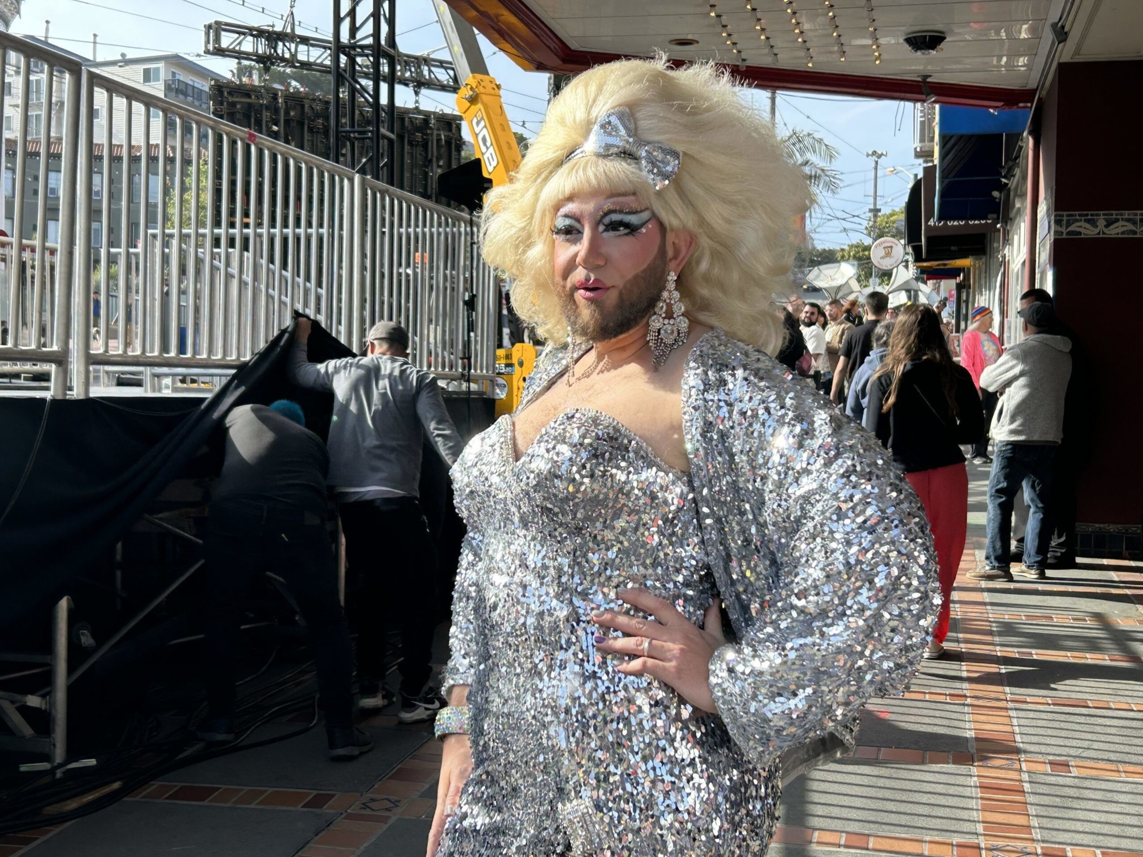 A person dressed in a sparkling silver outfit and big blonde wig poses confidently on a sunny urban sidewalk with people and scaffolding behind them.