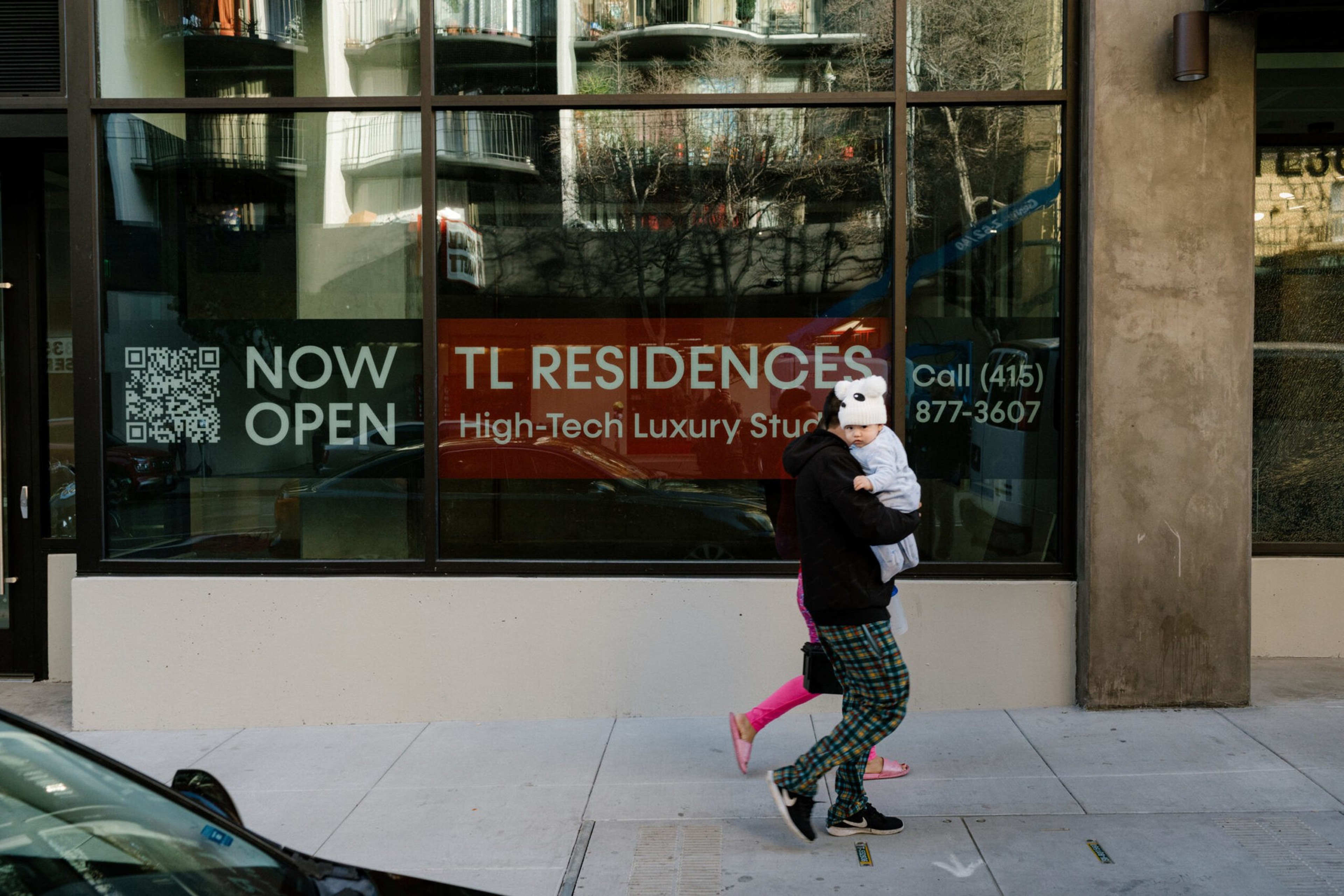 A person wearing plaid pants carries a baby in front of a window advertising "TL Residences" with "Now Open" and a phone number.