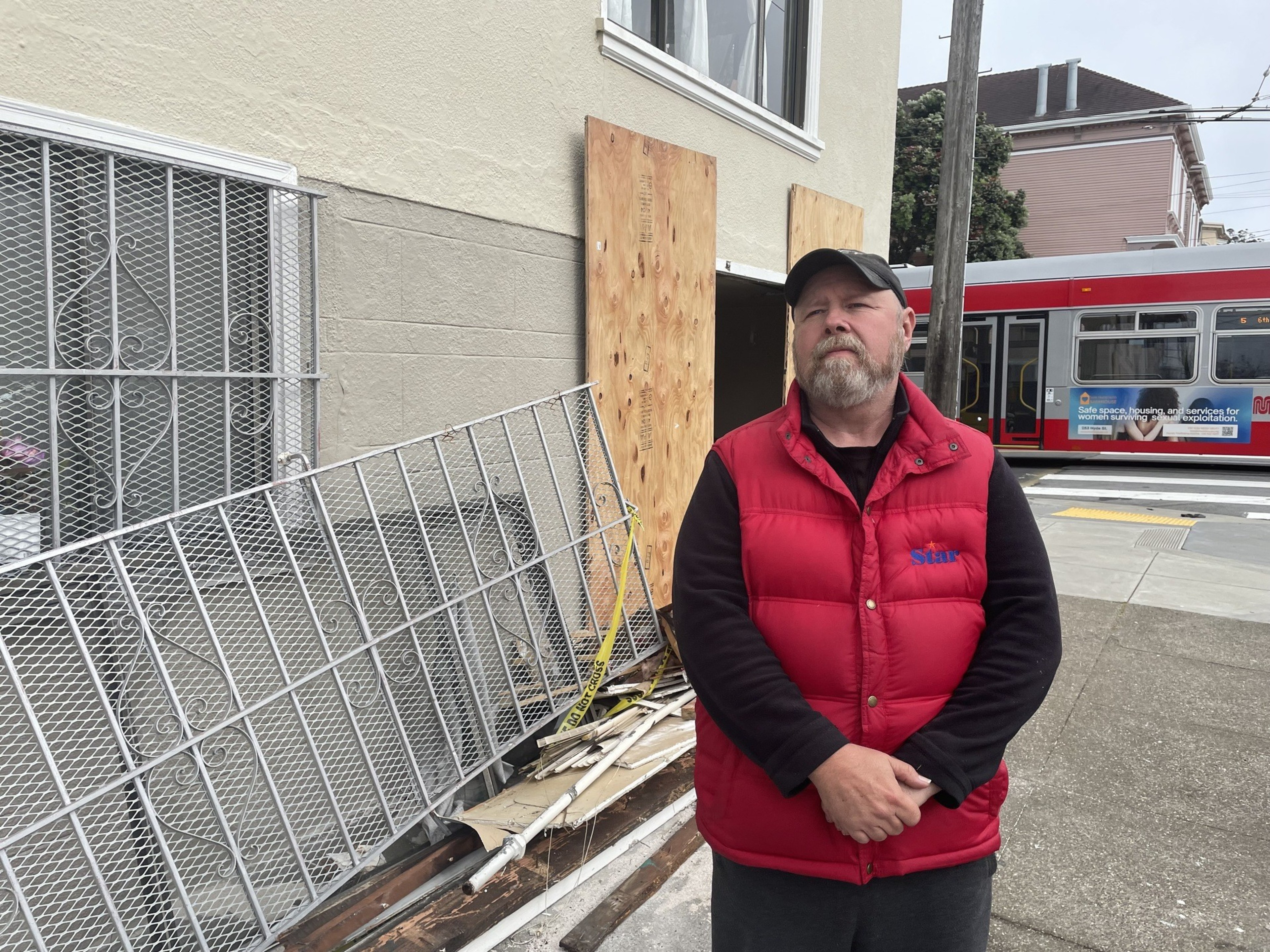 A man in a red vest stands in front of a building with boarded-up windows, near a metal fence and red trolley bus in the background.