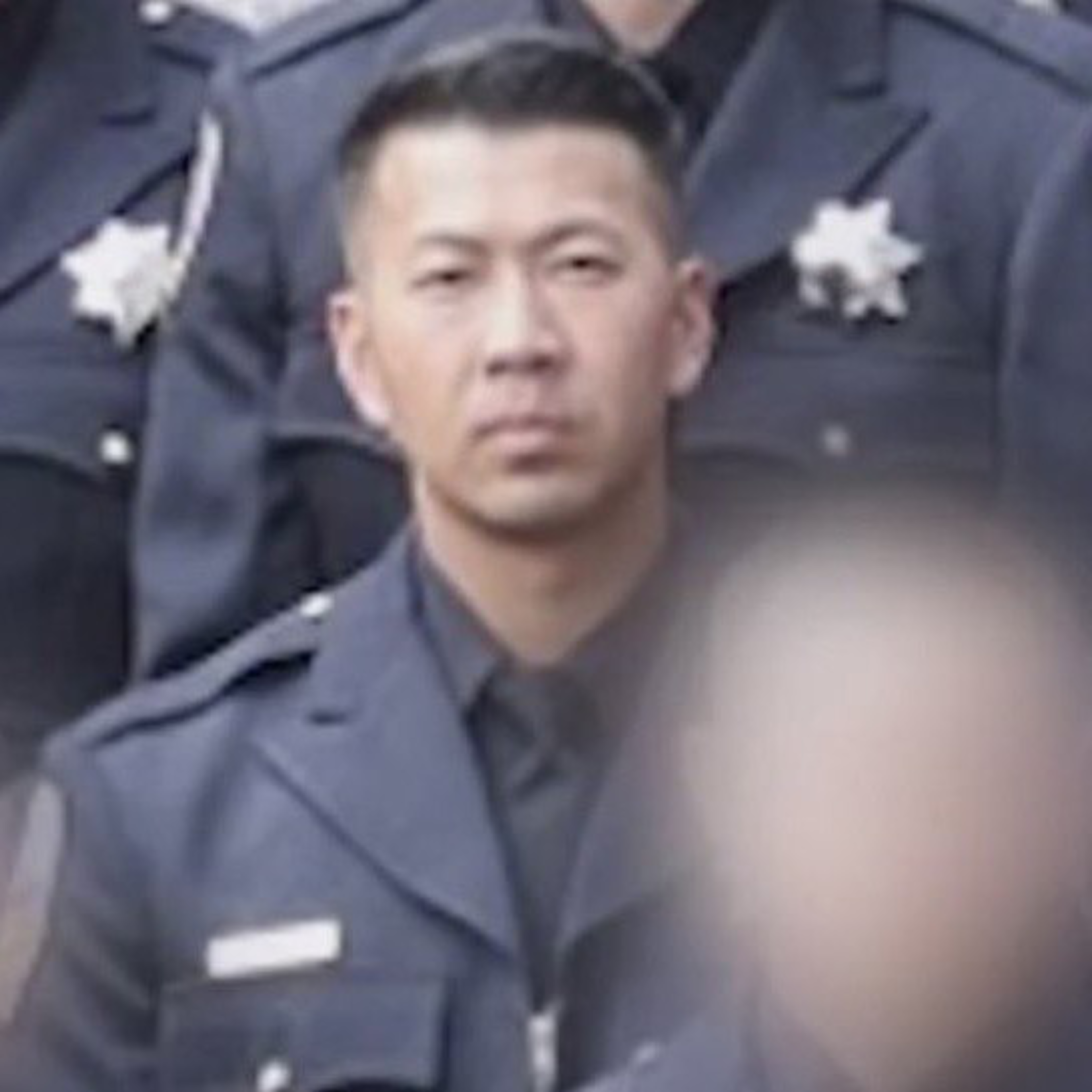 A person wearing a dark uniform with a badge on the chest stands among others in similar attire. The background is slightly blurred, focusing on the central figure.