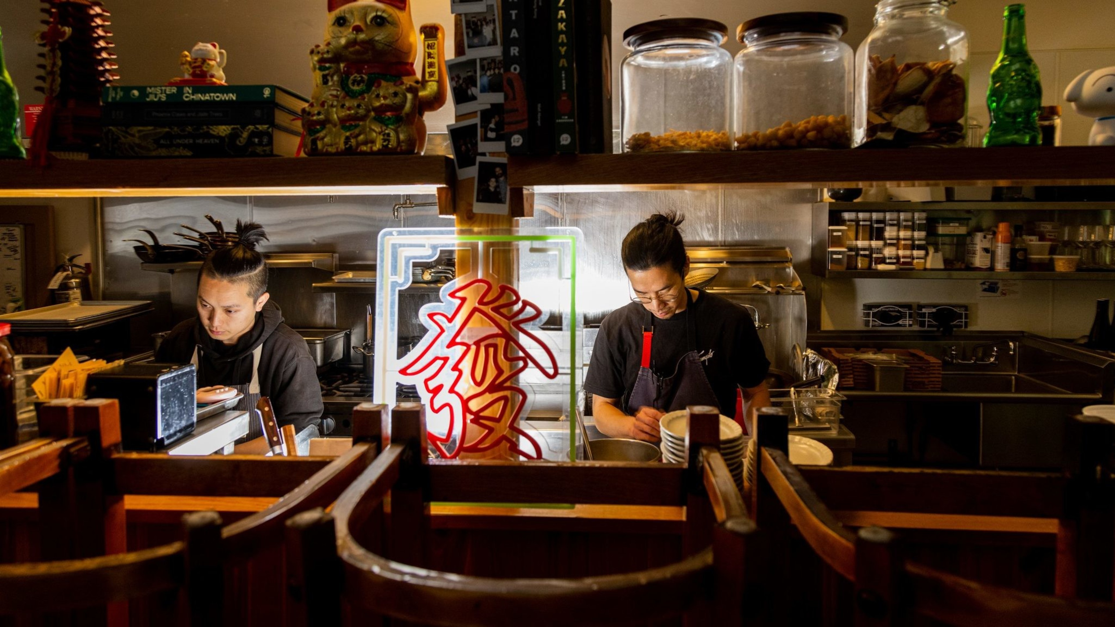 Two chefs work in a dimly lit kitchen, surrounded by jars and a neon sign with red characters. Shelves hold decorative items, including a waving cat.