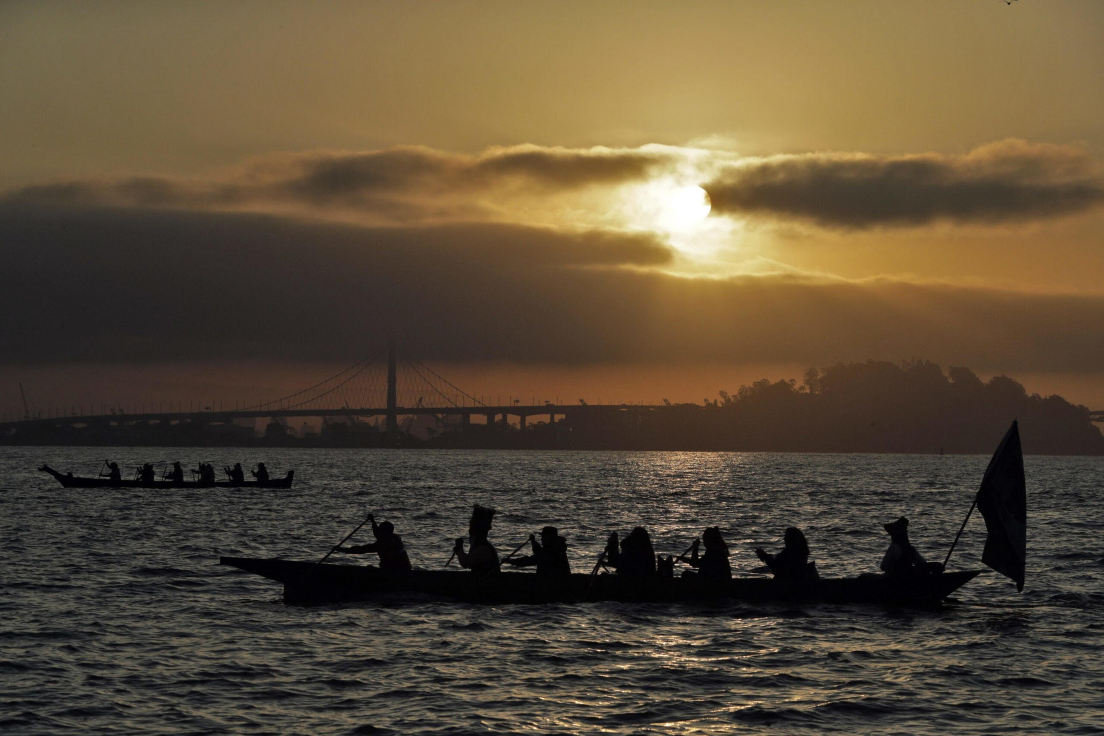 Eight people paddle in a long canoe on water during sunset with a bridge and land silhouetted against the sky in the background.