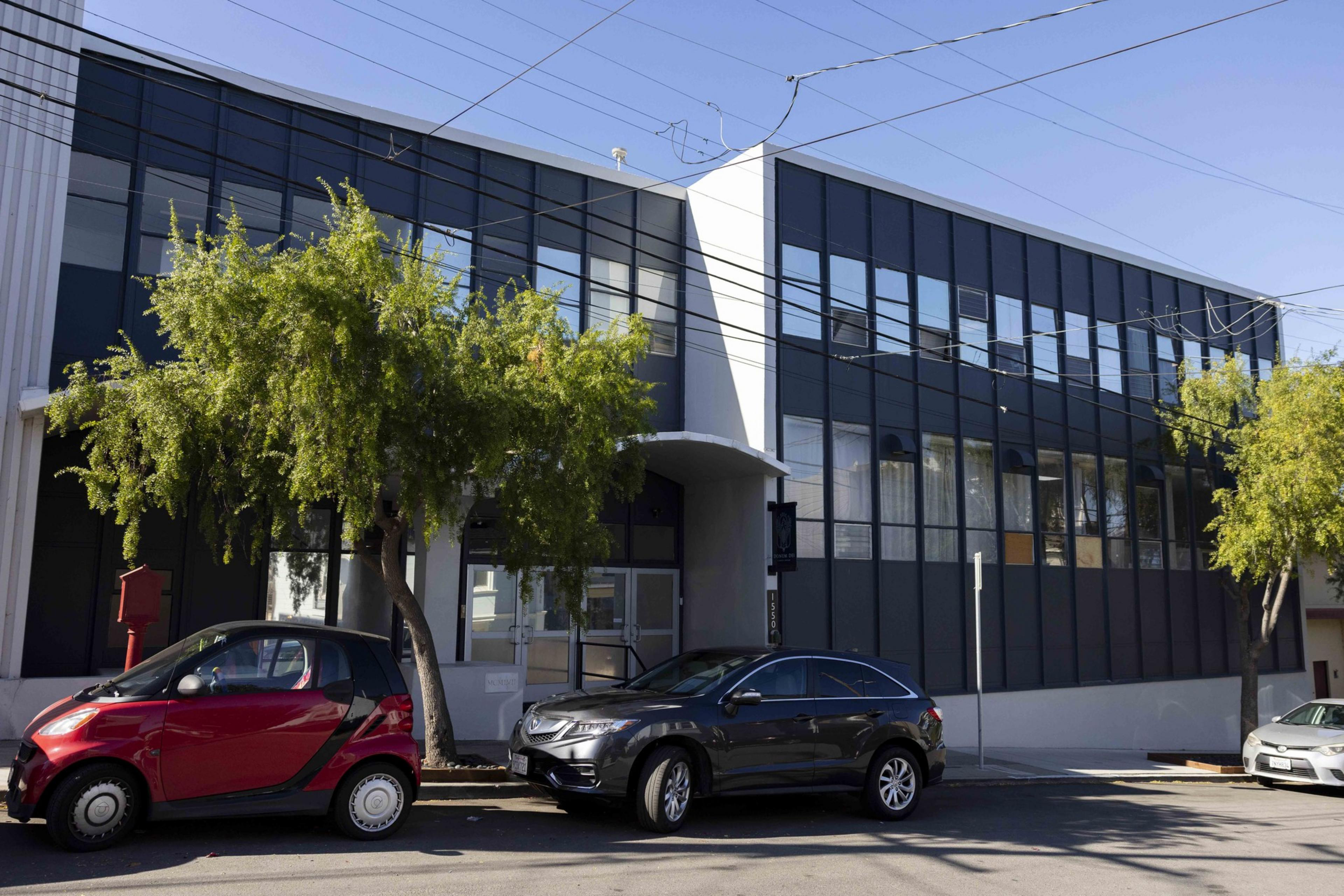 A modern two-story building with large windows is bordered by two trees. Two cars, one red and one gray, are parked in front on a sunny day.