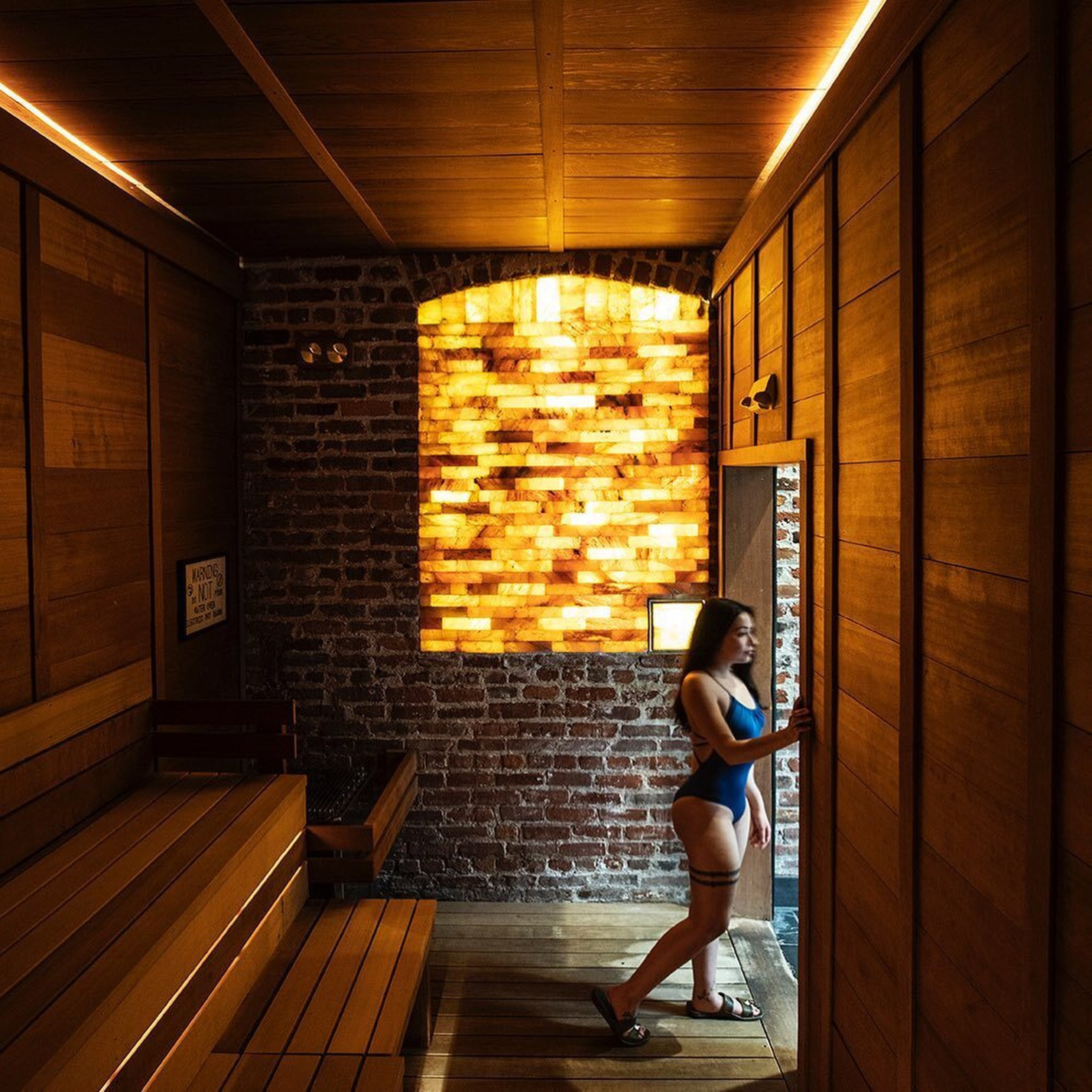 A woman in a swimsuit stands by the door of a cozy wooden sauna with glowing salt bricks, benches, and a sign on the brick wall.