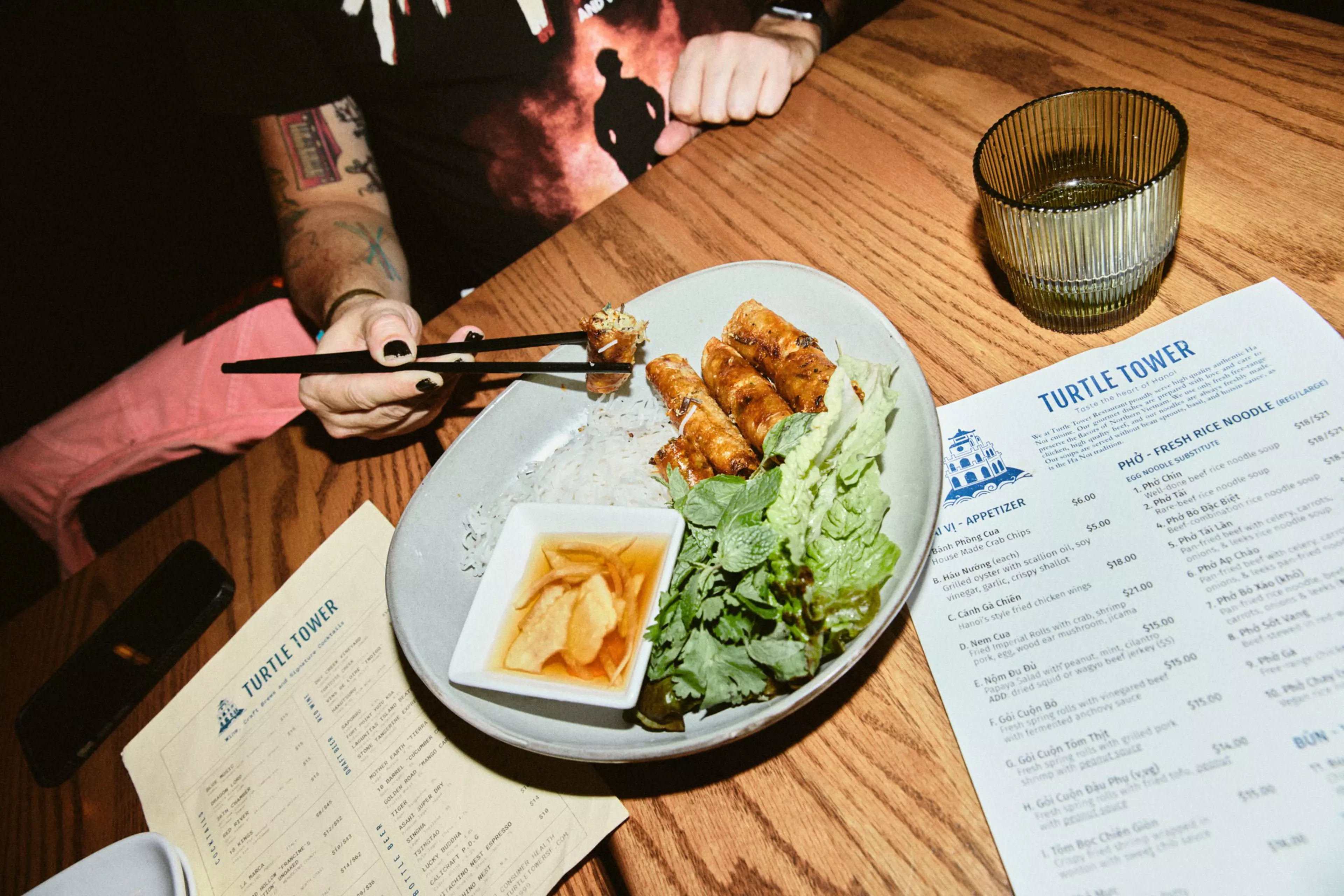 A person with tattoos holds chopsticks picking up spring rolls from a plate with rice, fresh herbs, lettuce, and a dipping sauce, next to menus and a glass.