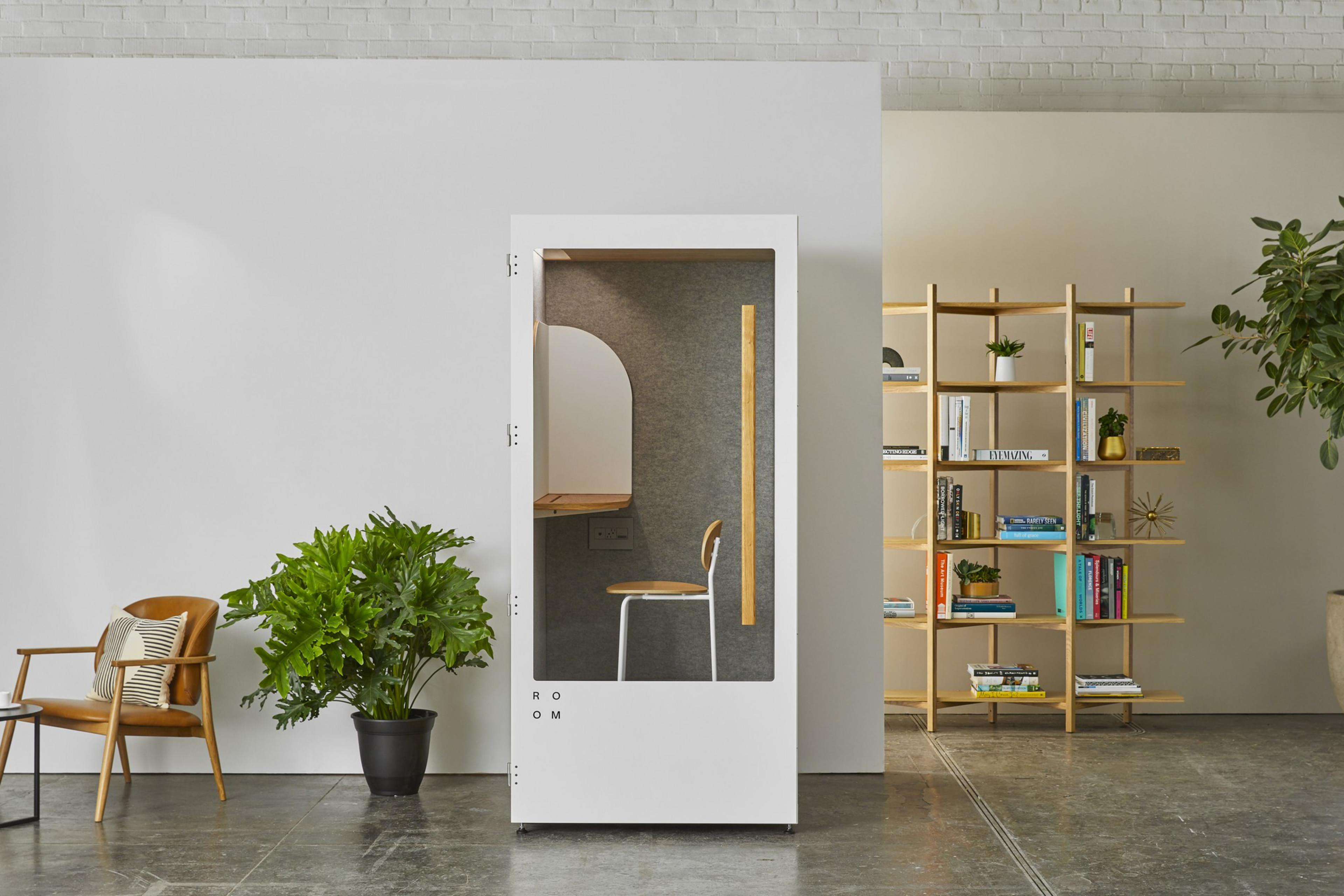 A Room phone booth sits center of frame; a bookshelf is in the background and a plant and a chair sit to the left of the booth