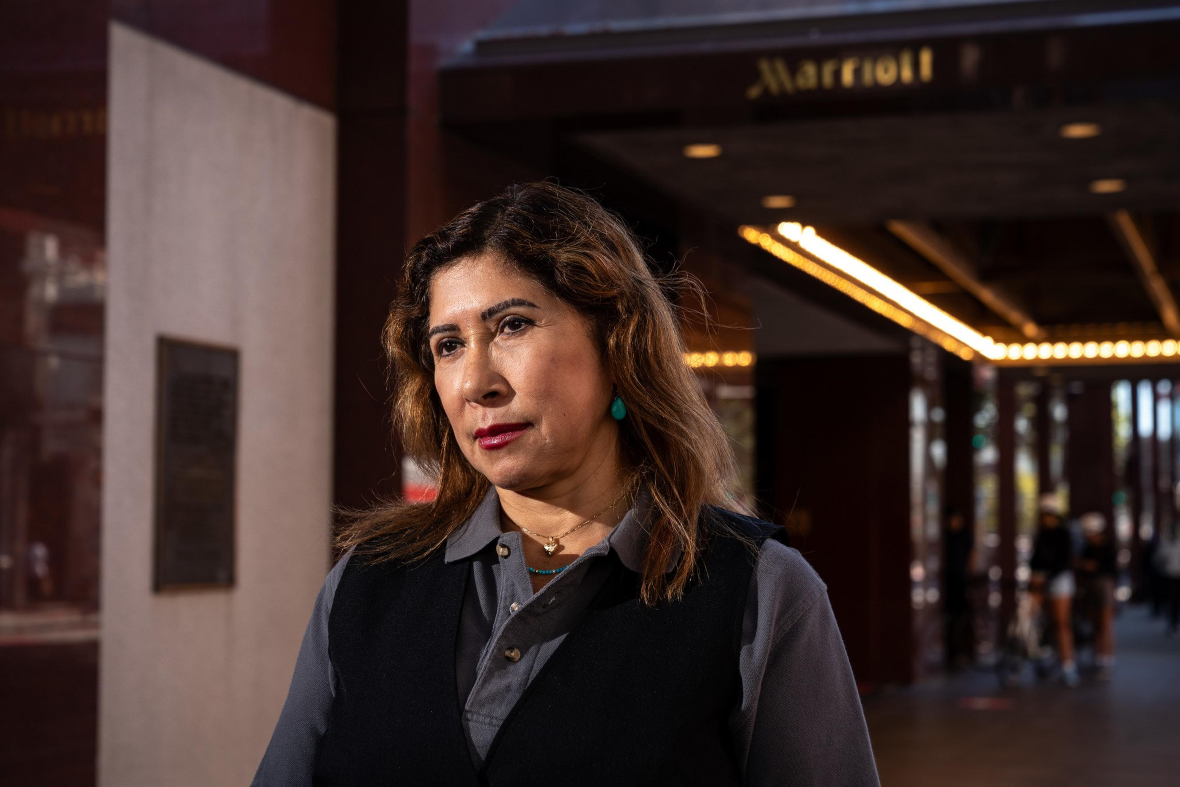 A woman stands in front of a Marriott hotel entrance. She has shoulder-length hair, wears a black vest over a gray shirt, and looks contemplatively to the side.