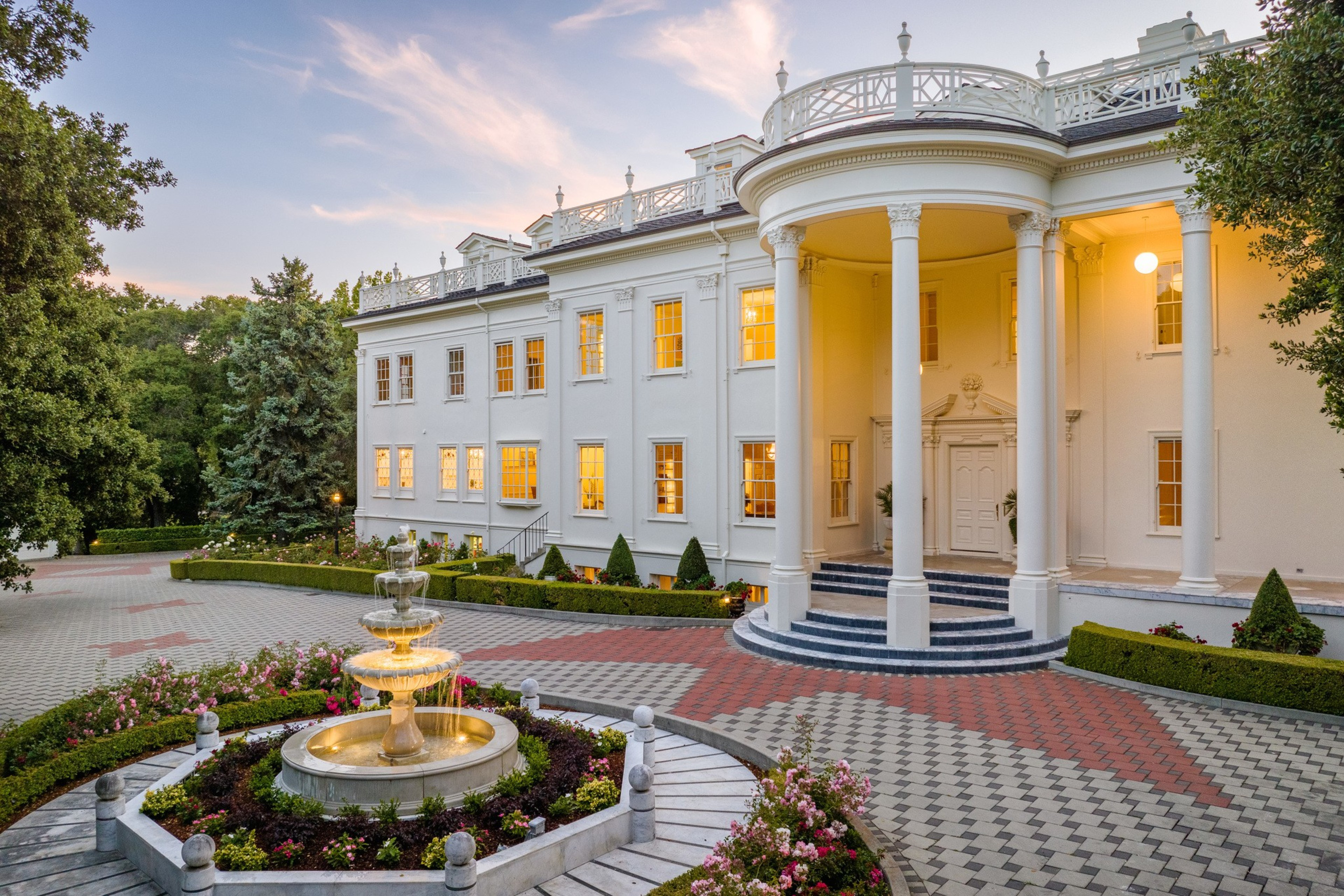 An ornate circular fountain burbles in front of a large white building at evening time