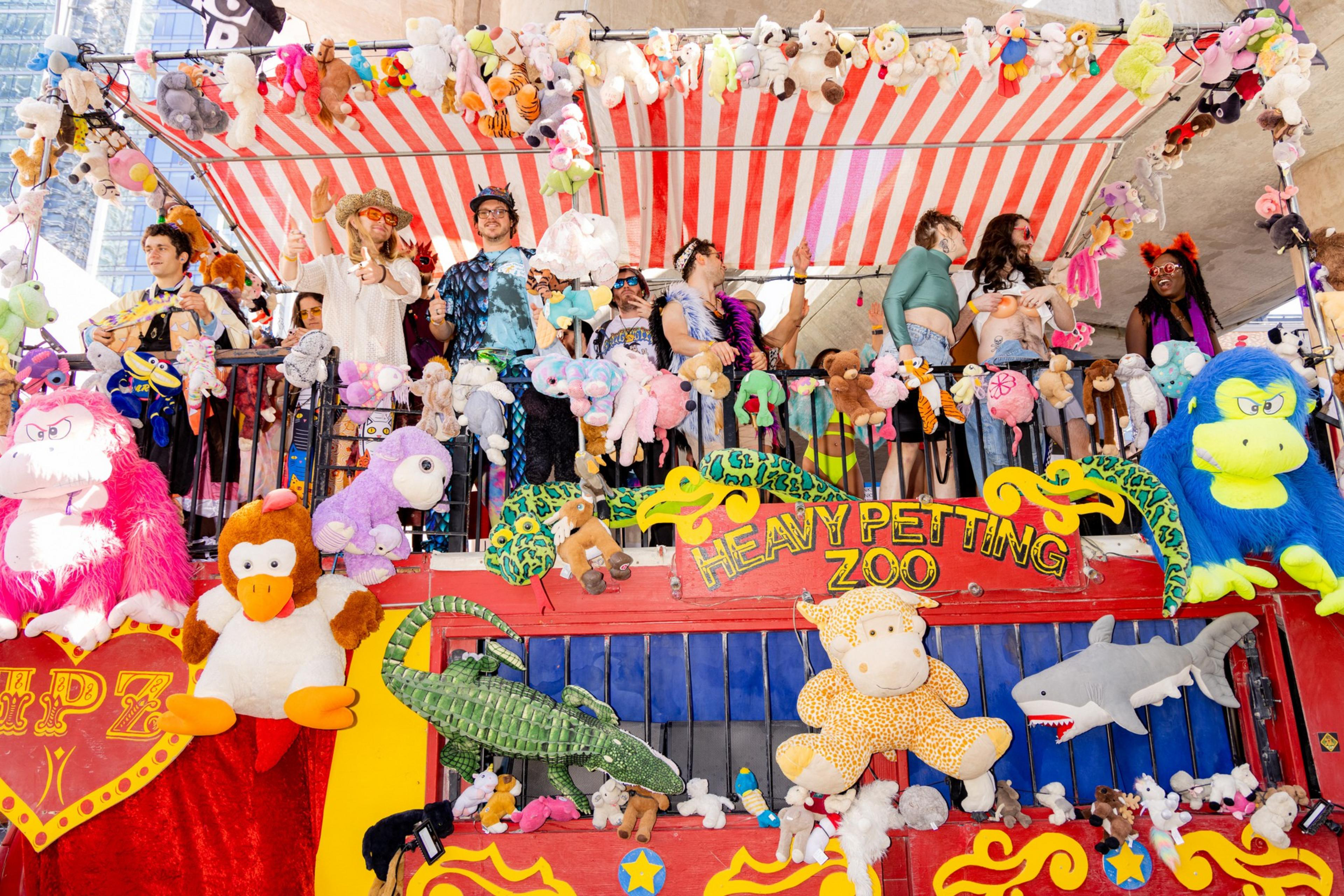 A lively group of people stands on a brightly decorated parade float adorned with stuffed animals and a striped canopy, featuring a &quot;Heavy Petting Zoo&quot; sign.