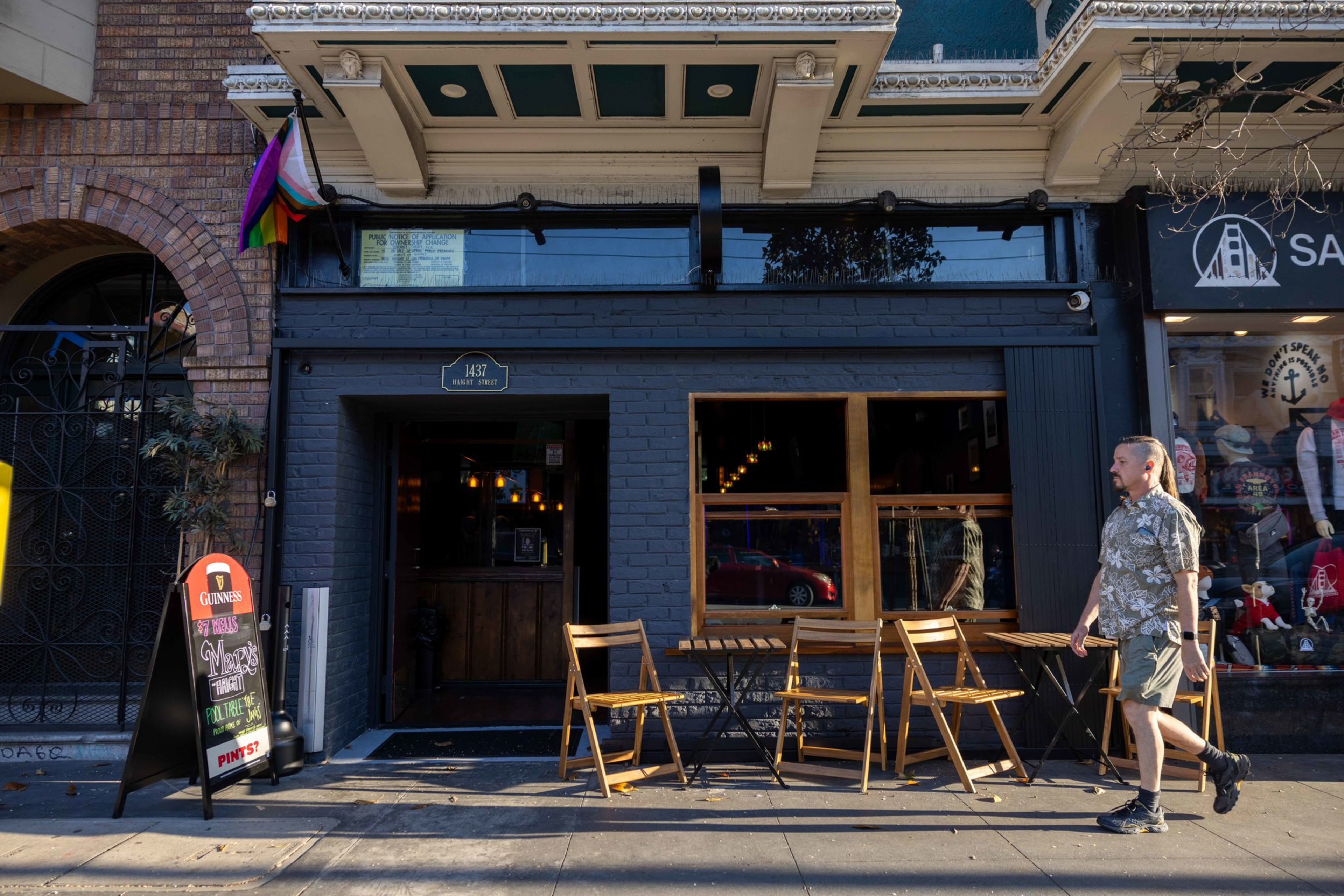 A man walks past a dark-blue brick bar with wooden tables and chairs outside, a pride flag above the entrance, and a sandwich board sign on the sidewalk.