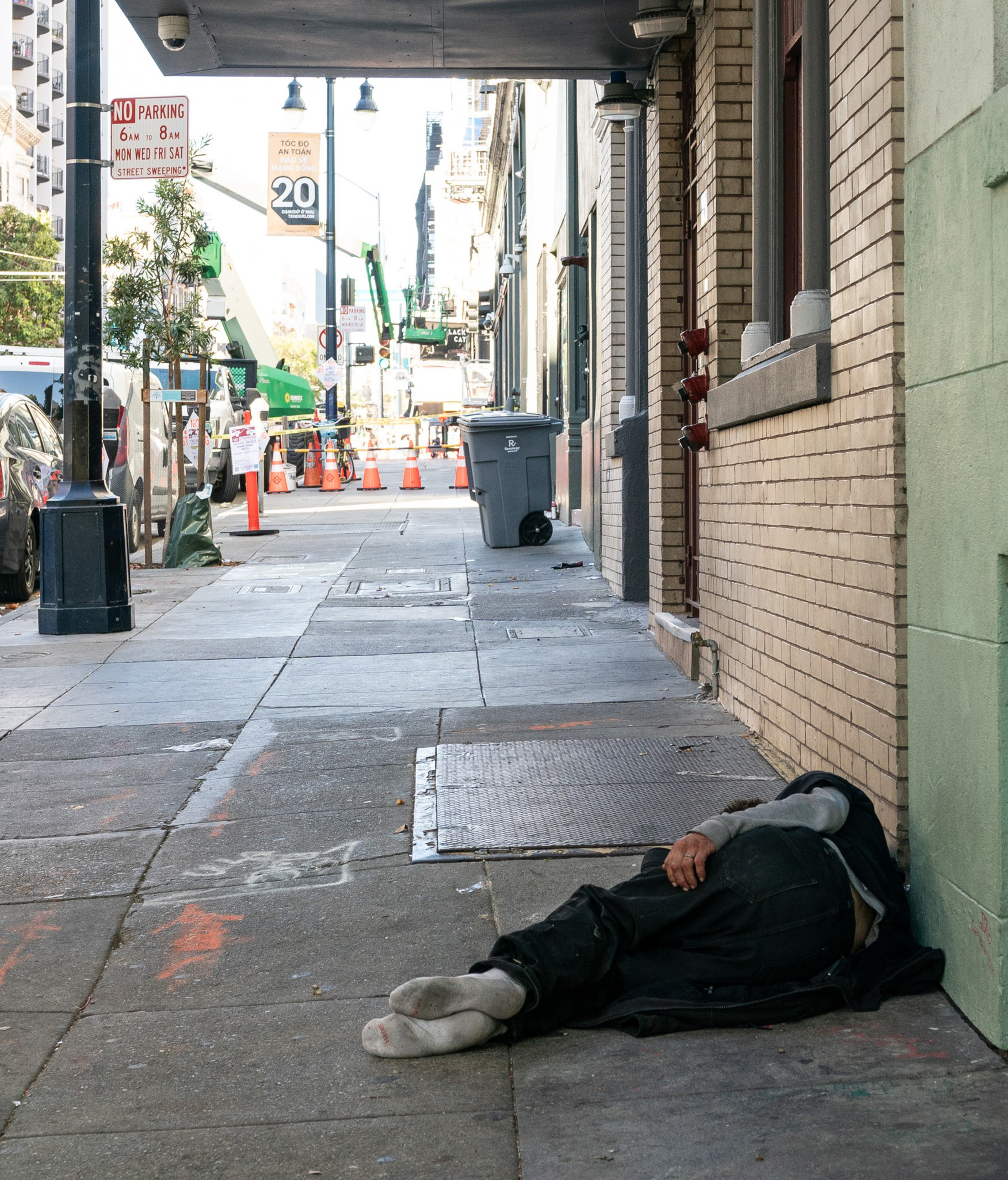 A person is lying on a sidewalk by a building, with traffic cones and vehicles visible in the street in the background.