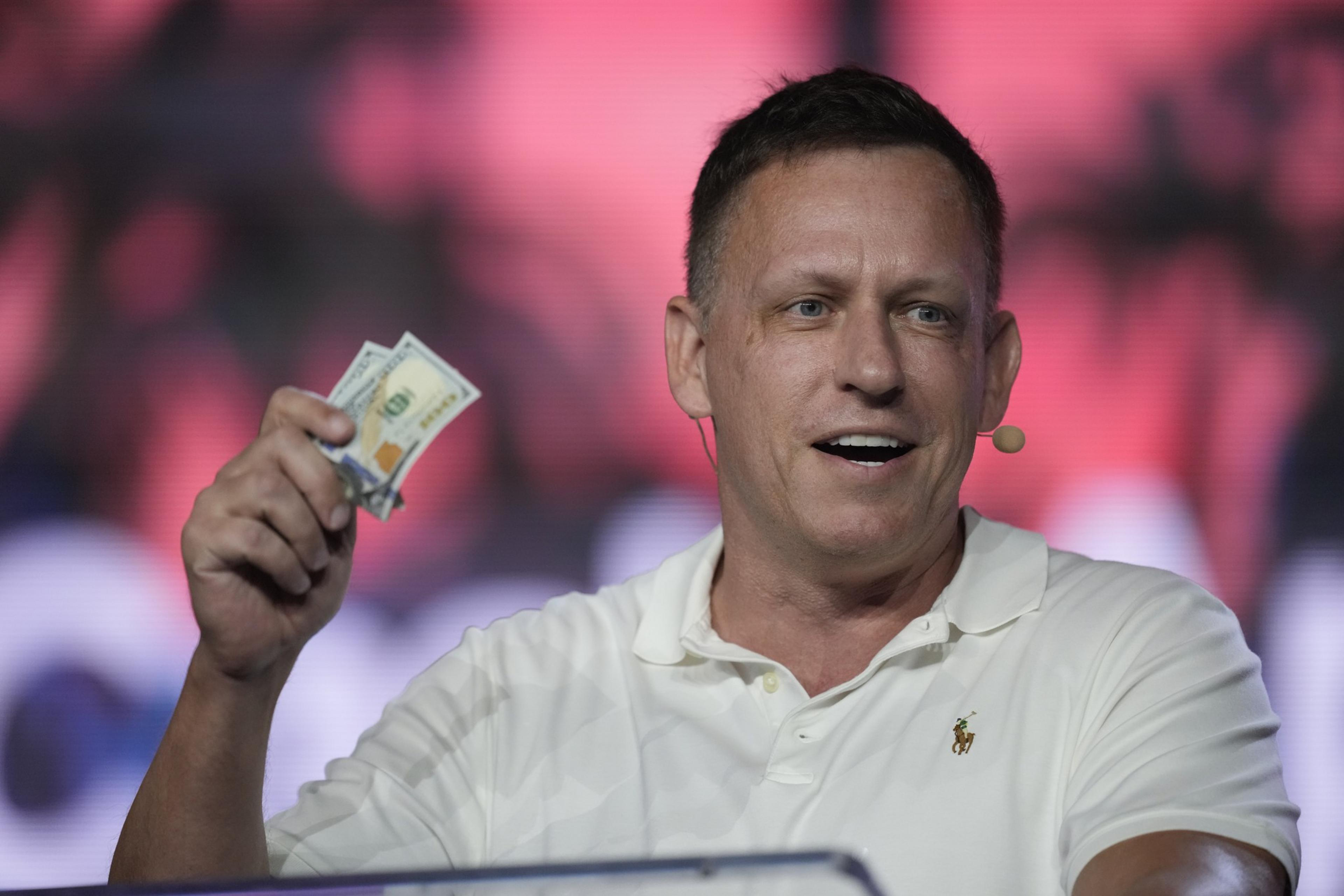 A man wearing a white polo shirt and a headset microphone holds up two $100 bills with a smile against a blurred red and black background.