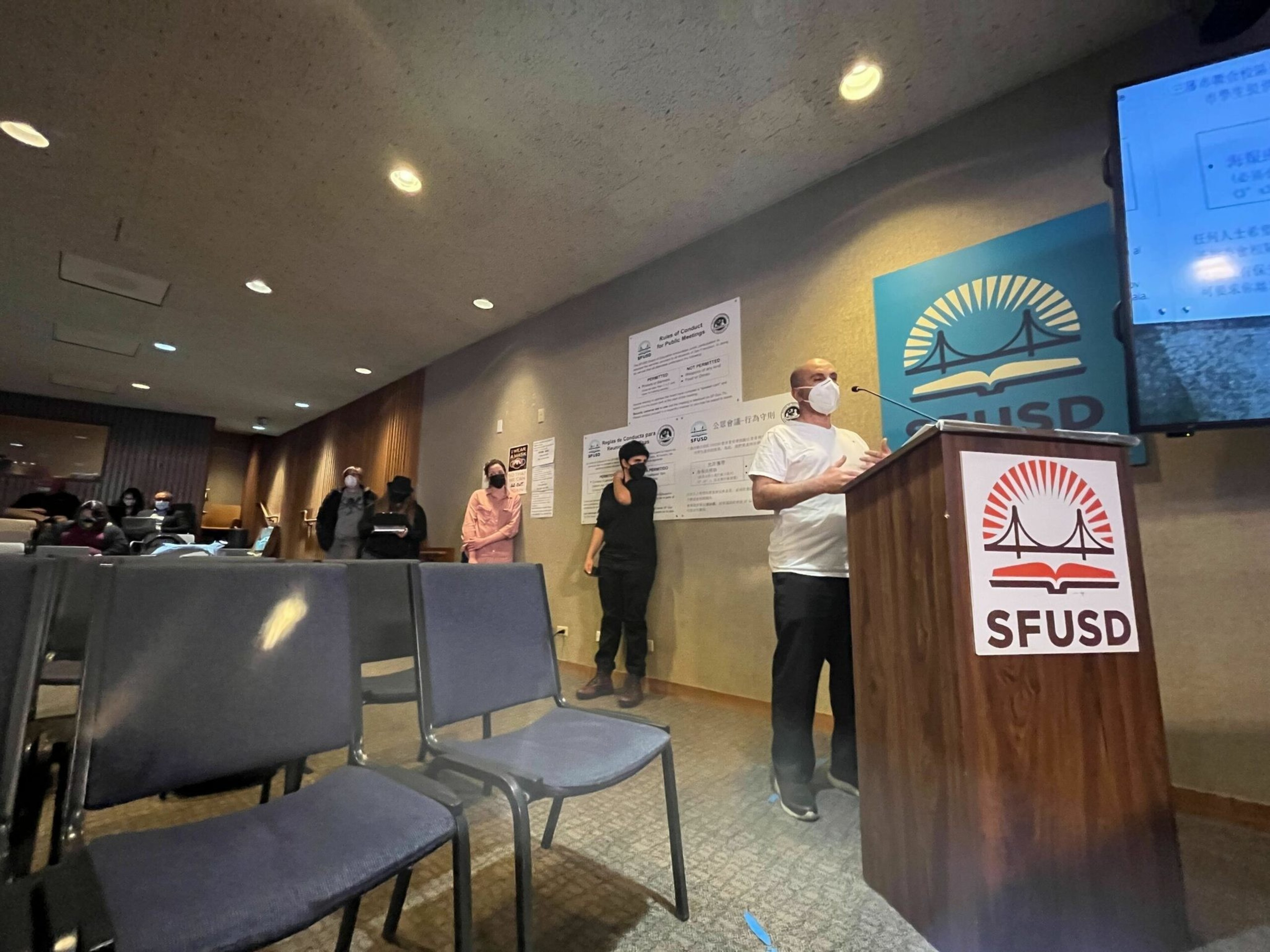 A man wearing a mask speaks at an SFUSD podium while several masked people stand in line against the wall in a dimly lit room with empty chairs.