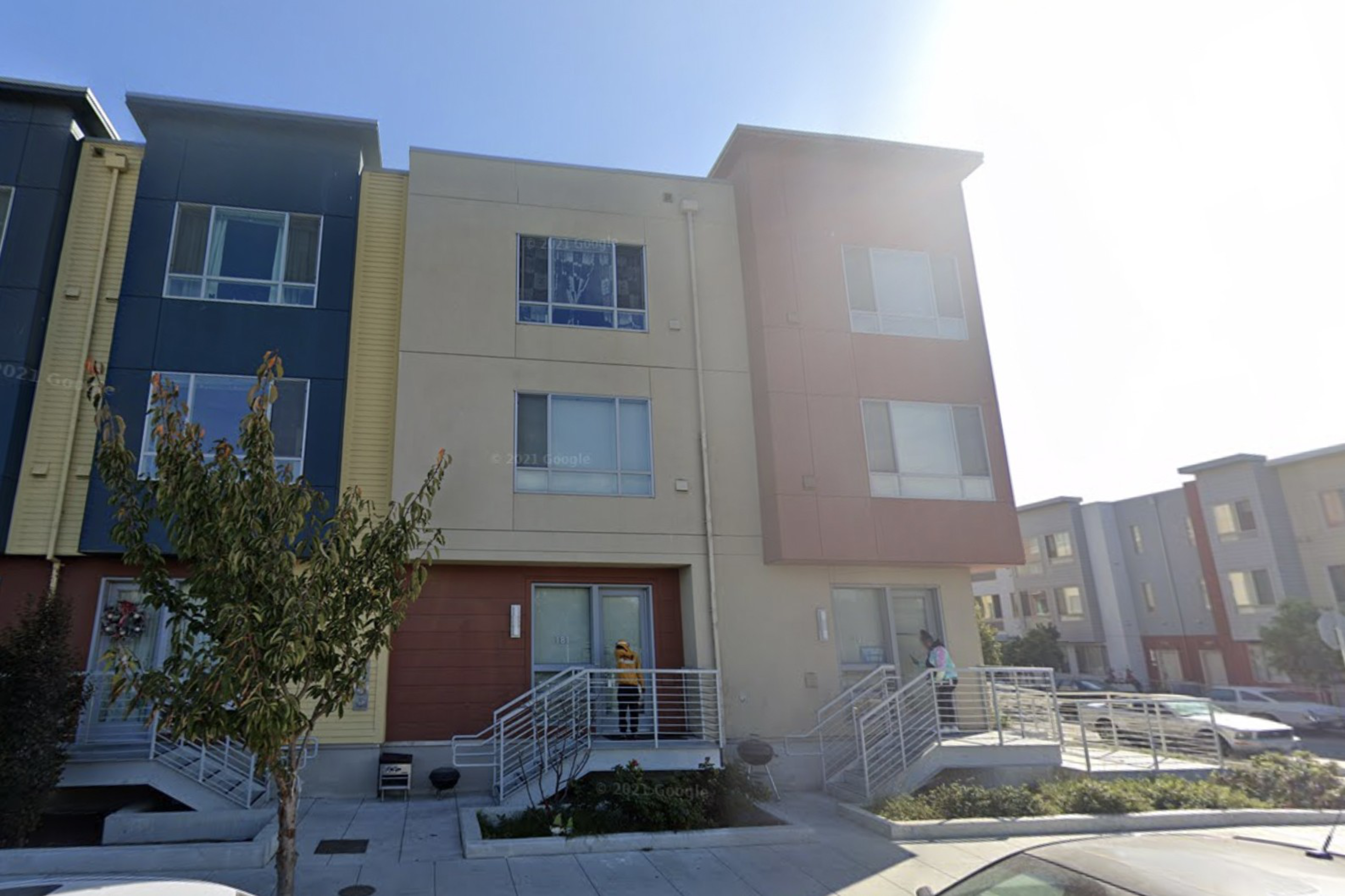 A row of modern three-story townhouses with large windows, each entry accessed by raised stairs with metal railings. A tree and parked cars are in front.