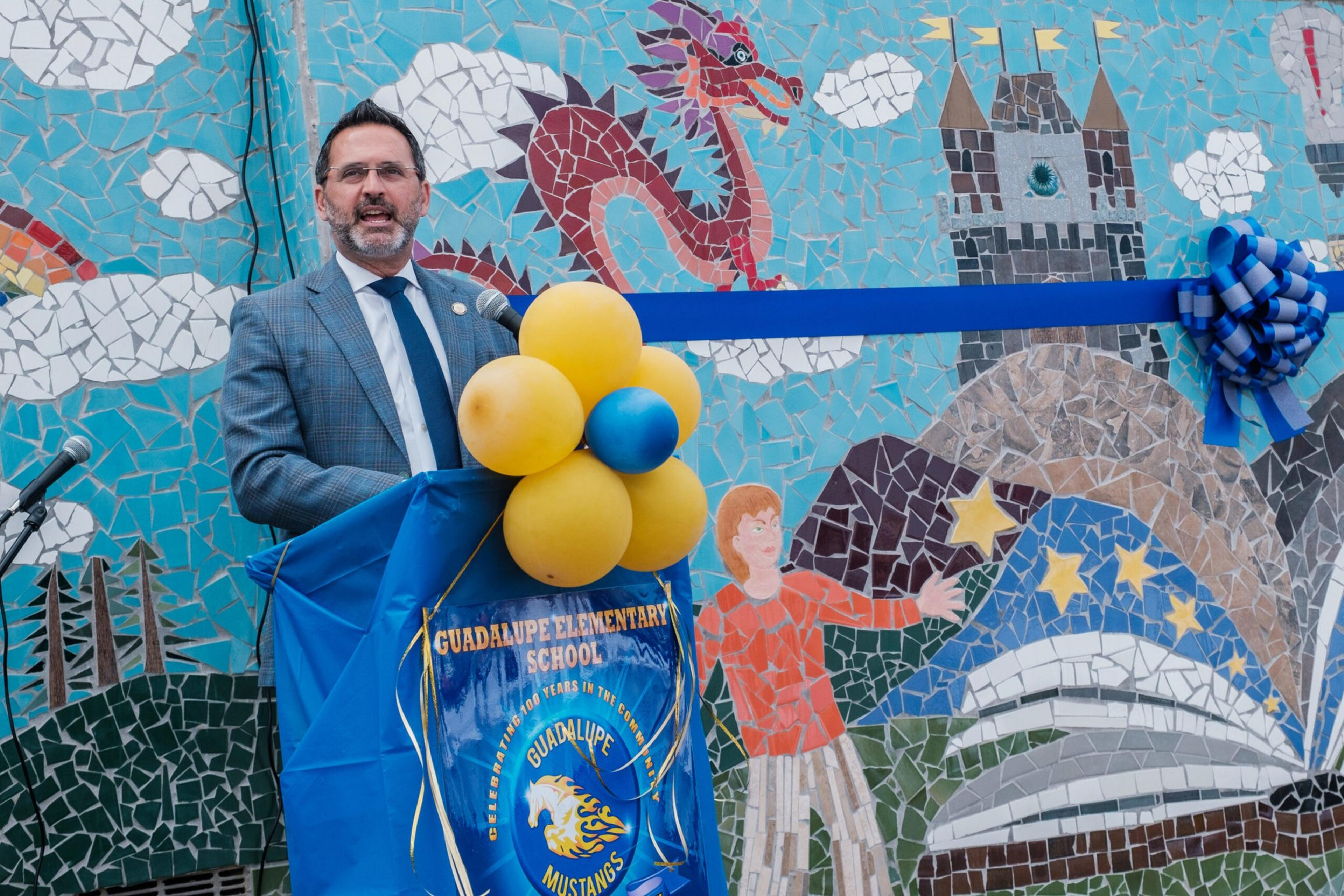 A man in a suit speaks at a podium decorated with yellow and blue balloons in front of a colorful mosaic mural featuring a red dragon, castle, and a child reaching for stars.