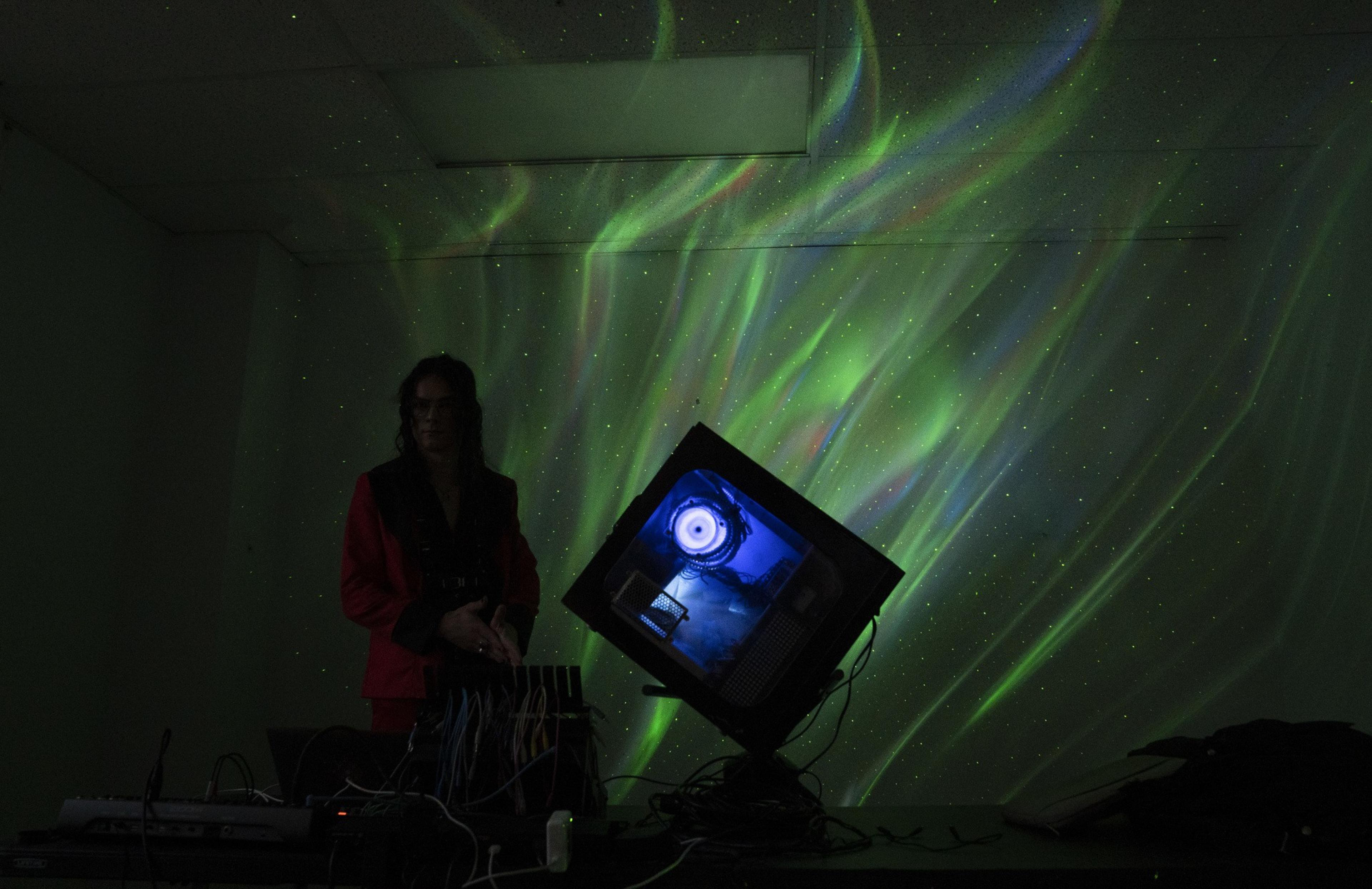 A person stands in a dark room with a colorful aurora-like light display on the wall. A tilted monitor on a table shows a circular, eye-like design.
