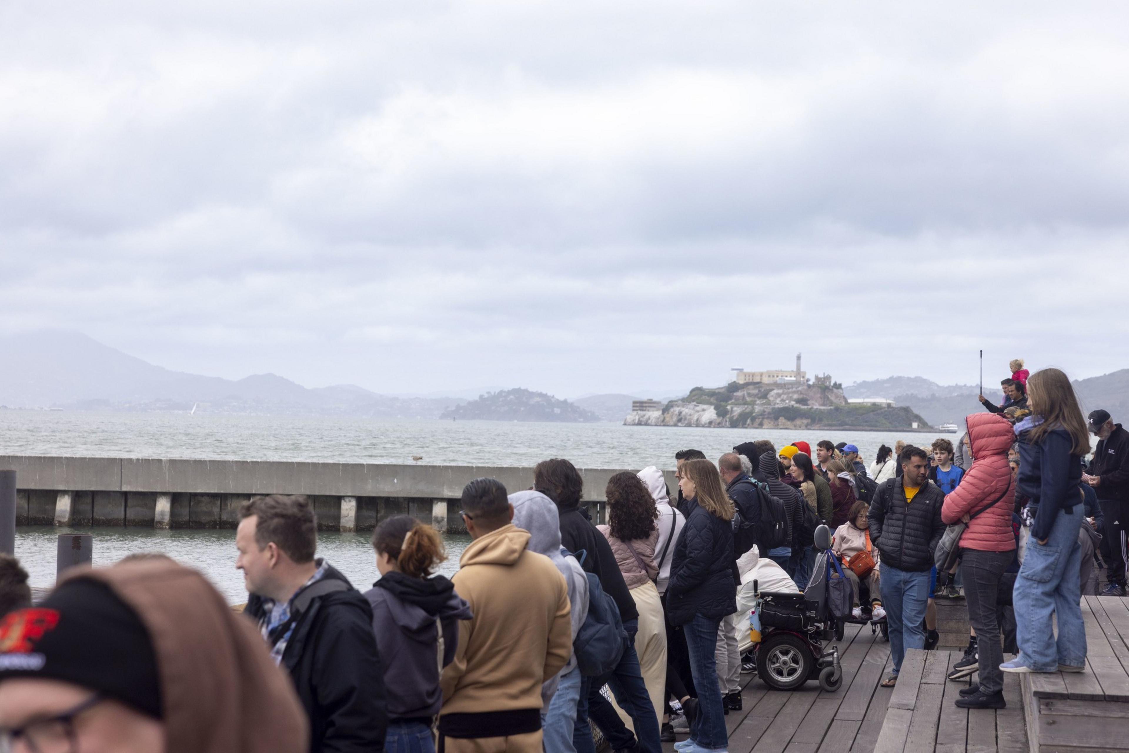 A crowd of people stands on a wooden pier, looking out over the water toward an island with a large building. The sky is cloudy, and some people wear jackets.