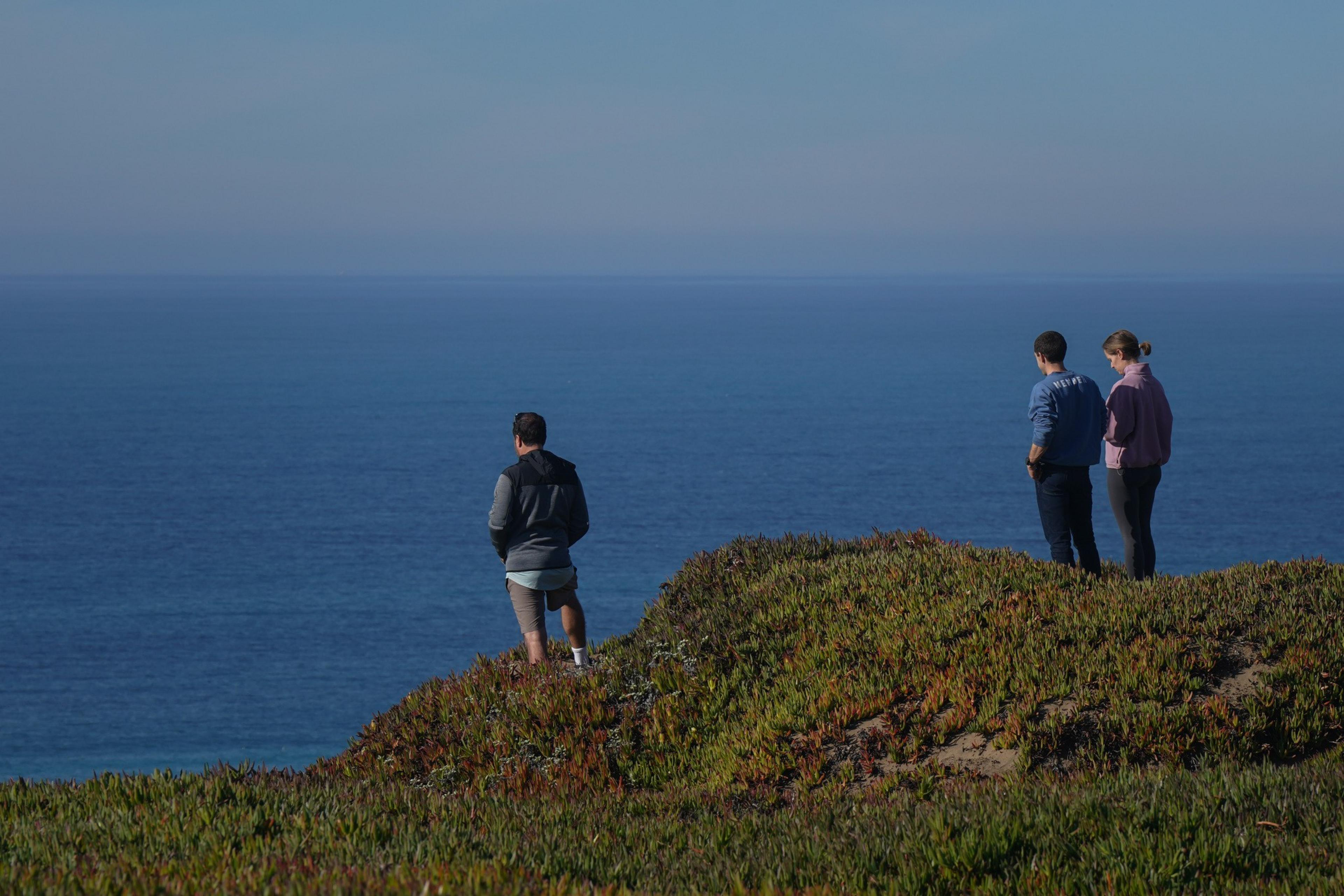 Three people stand on a grassy cliff, looking out at a vast, calm ocean under a clear blue sky.