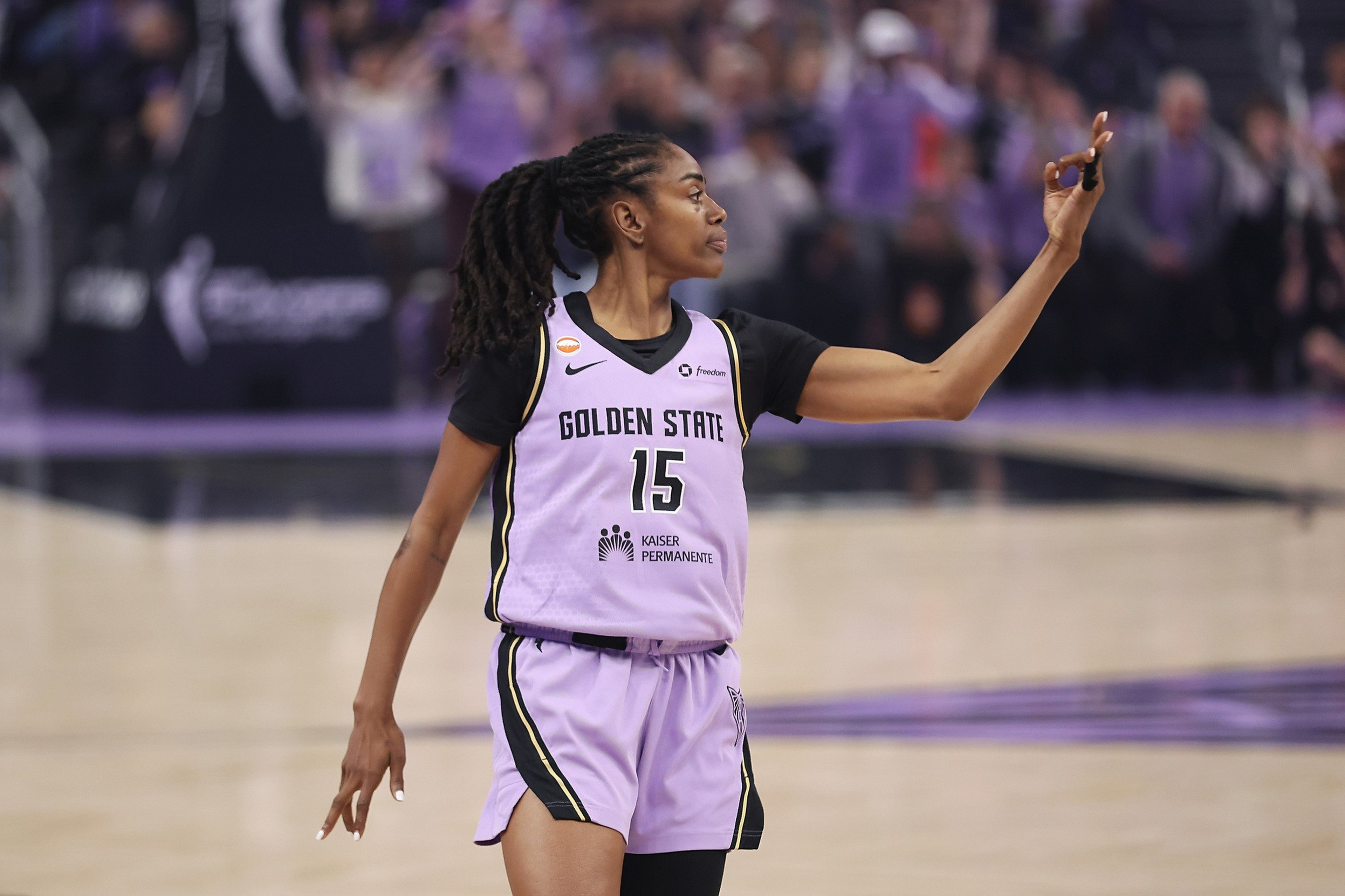 A basketball player wearing a Golden State jersey with number 15 signals with her right hand while walking on the court.