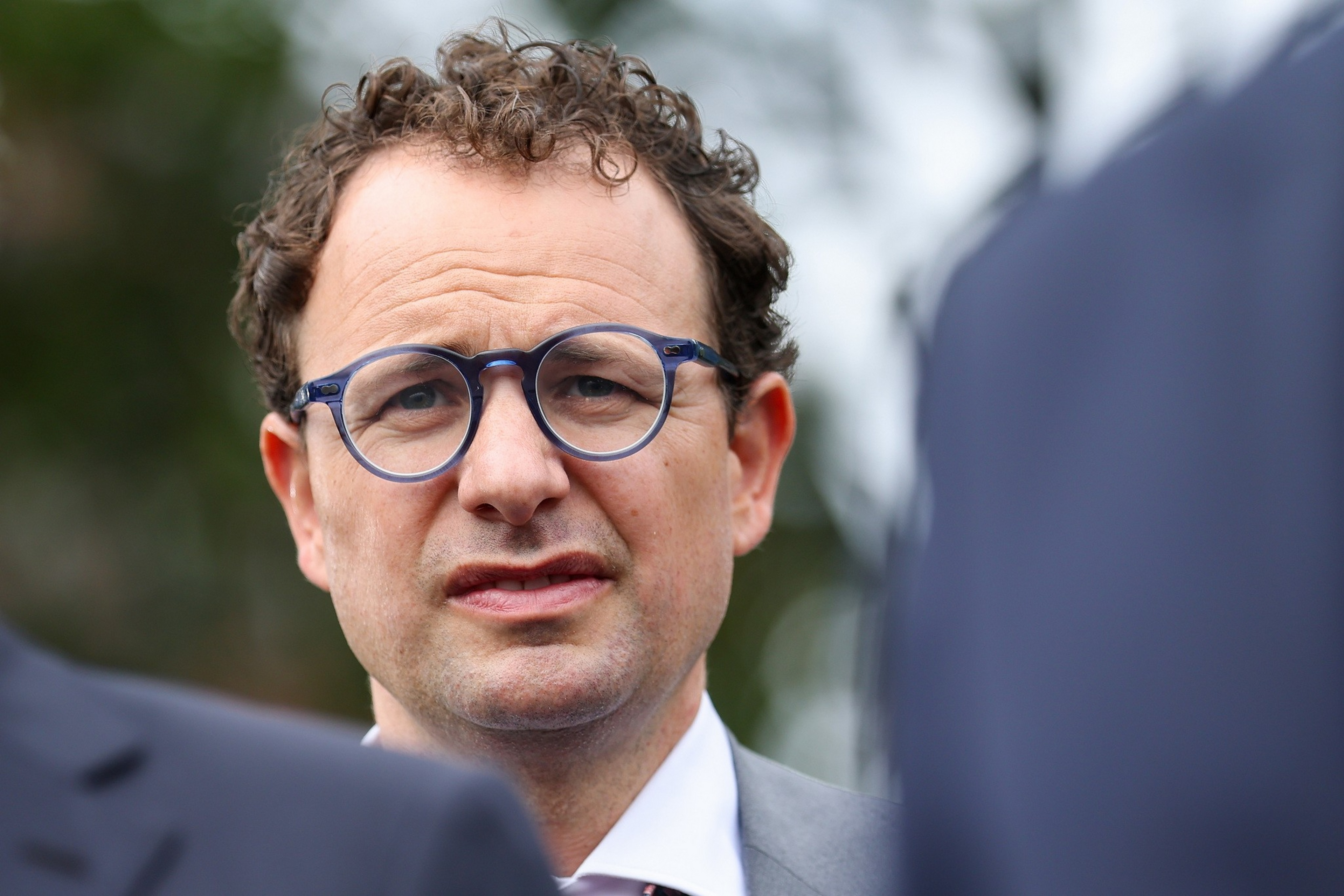 A man with curly brown hair and blue glasses frowns slightly, wearing a gray suit and white shirt, outdoors with blurred greenery and people nearby.
