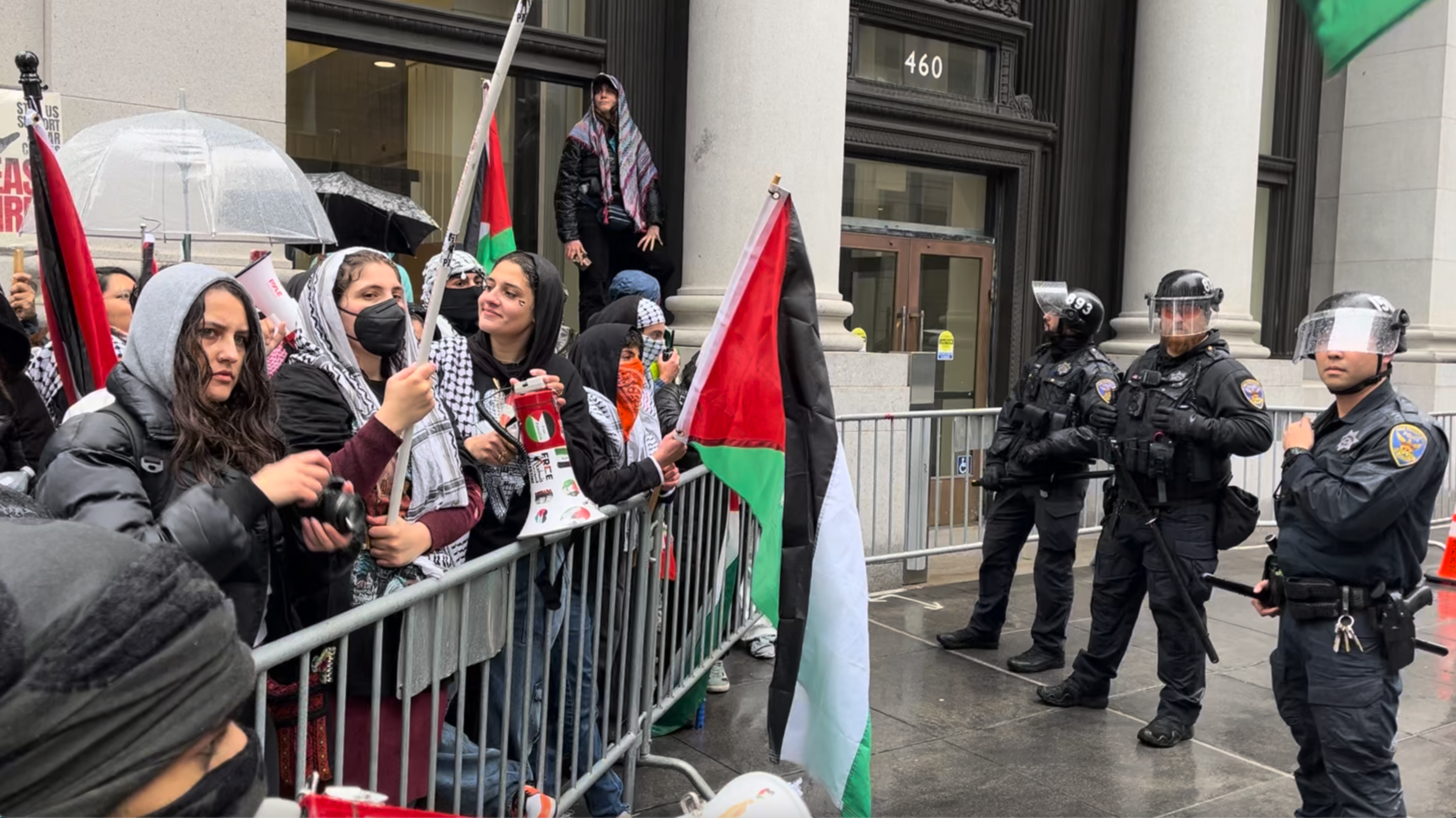A dozen protesters hold flags and stand behind one side of a metal barricade as three uniformed and helmeted police officers watch from the other side.