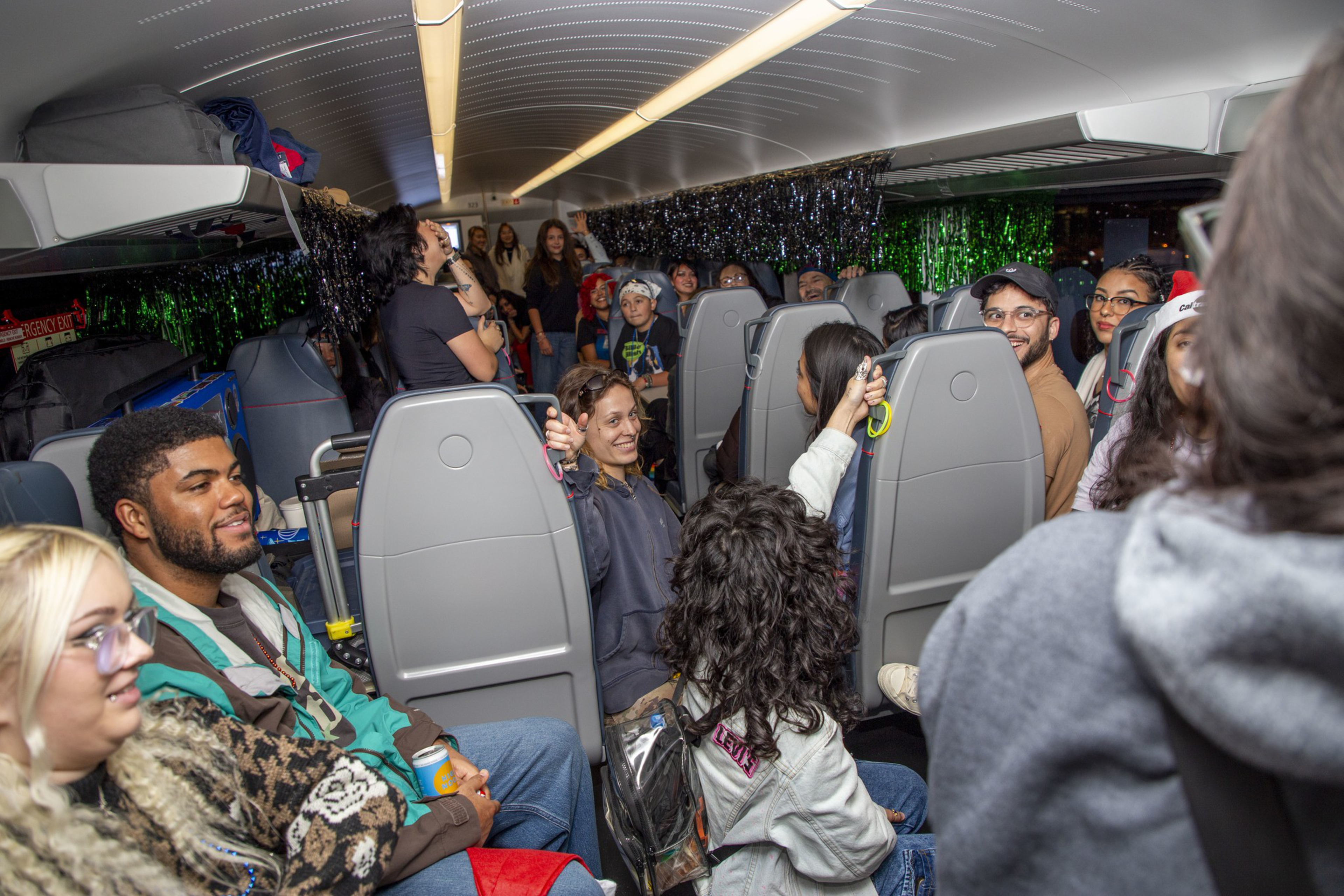 A group of people are sitting and standing inside a decorated train carriage, chatting and laughing. The shelves above hold bags, and the space feels lively and festive.